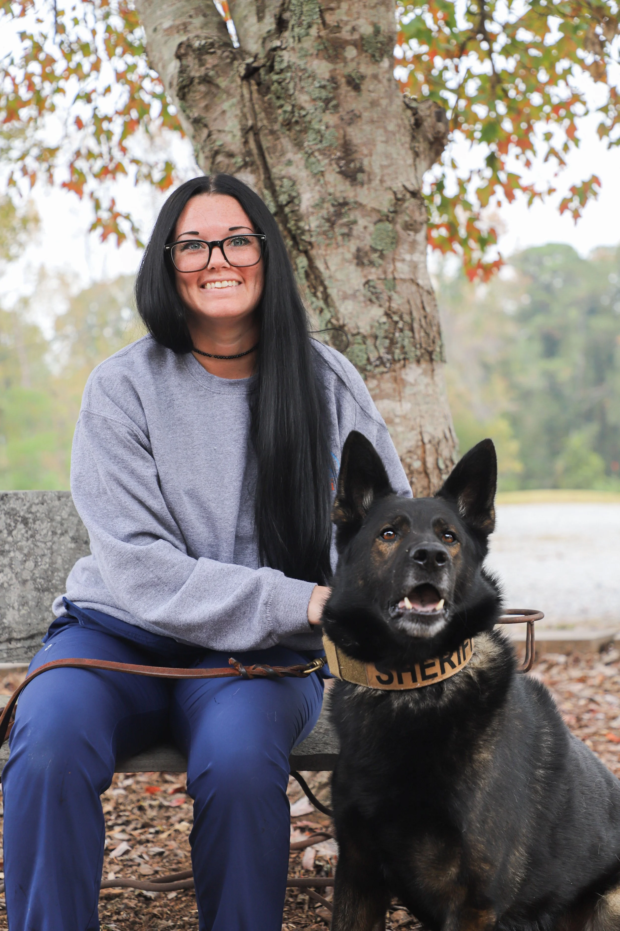 A woman with long black hair, glasses, and a gray sweatshirt sitting on a bench outdoors with a police dog, both smiling, in front of a large tree with fallen leaves.