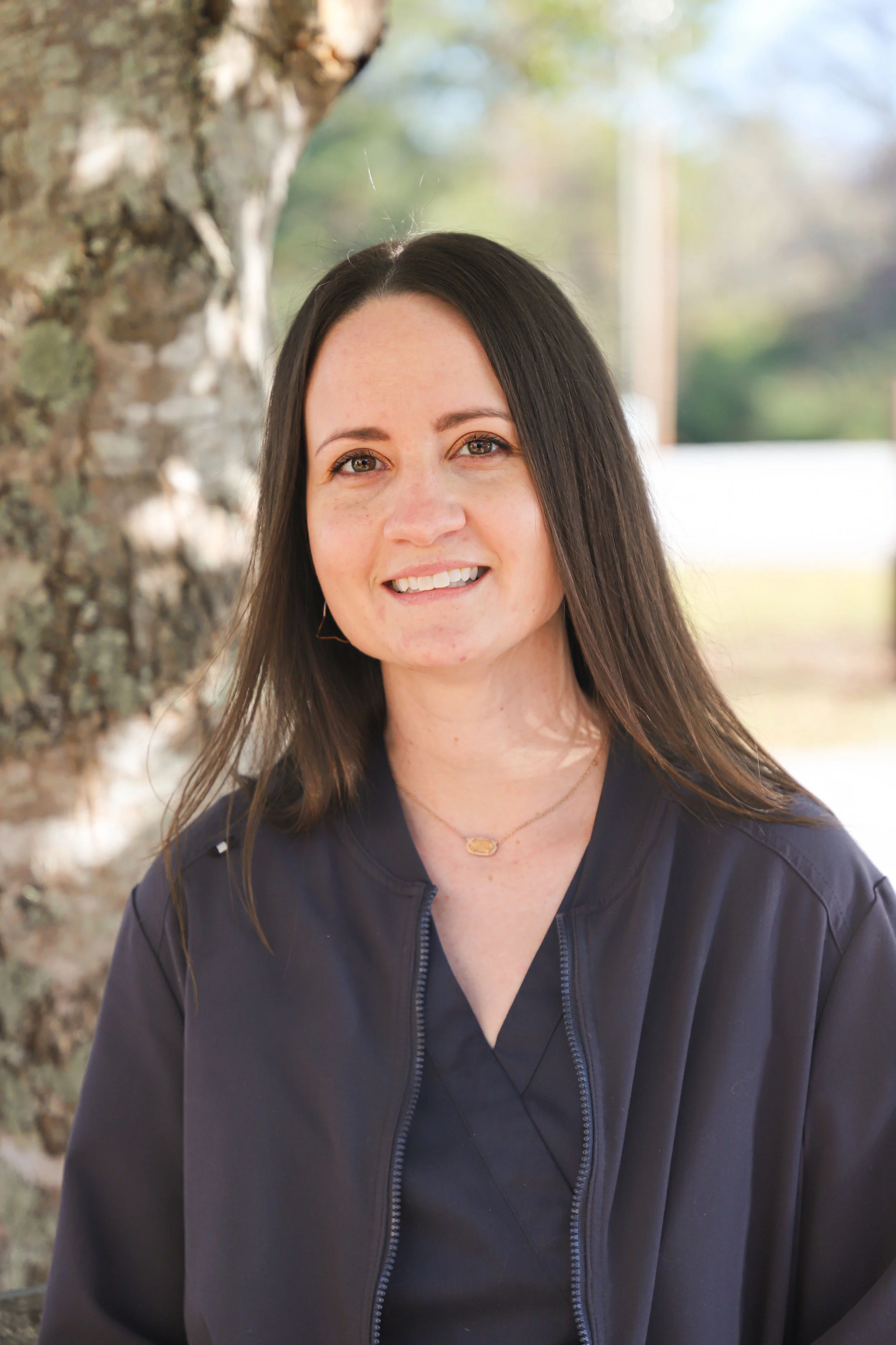A woman with shoulder-length dark hair smiling outdoors near a tree.