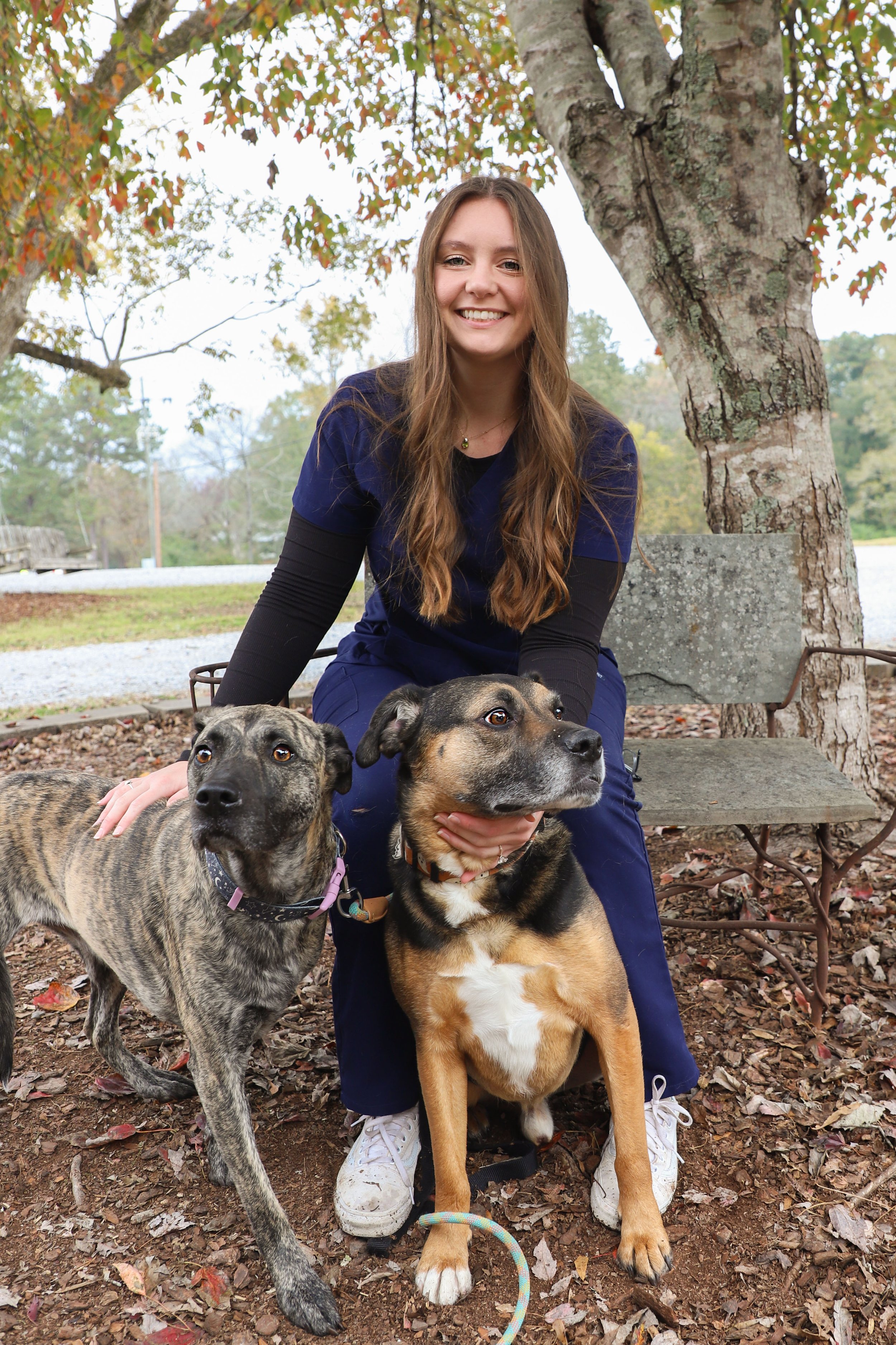 A woman in navy blue scrubs smiling and sitting outdoors on a wooden bench behind two dogs, with one dog sitting and the other standing, surrounded by fall leaves and trees.