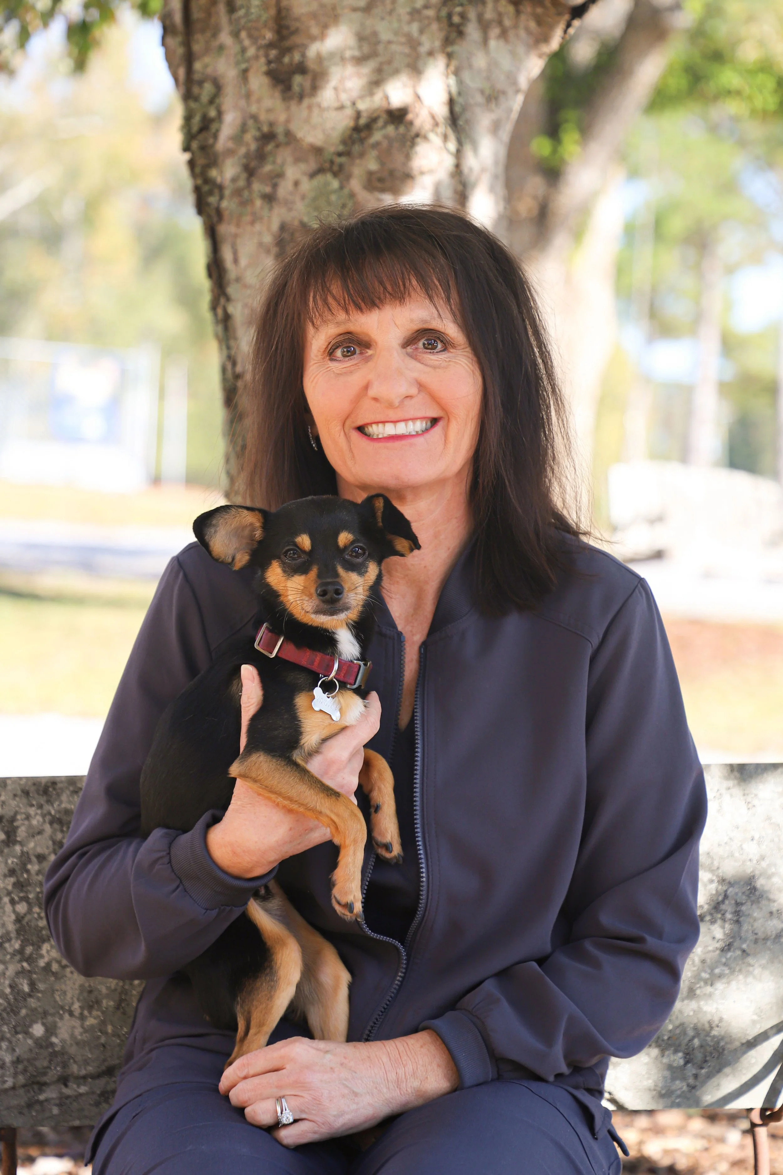 A woman with dark hair smiling and holding a small black and tan dog, sitting outdoors in front of a tree on a sunny day.