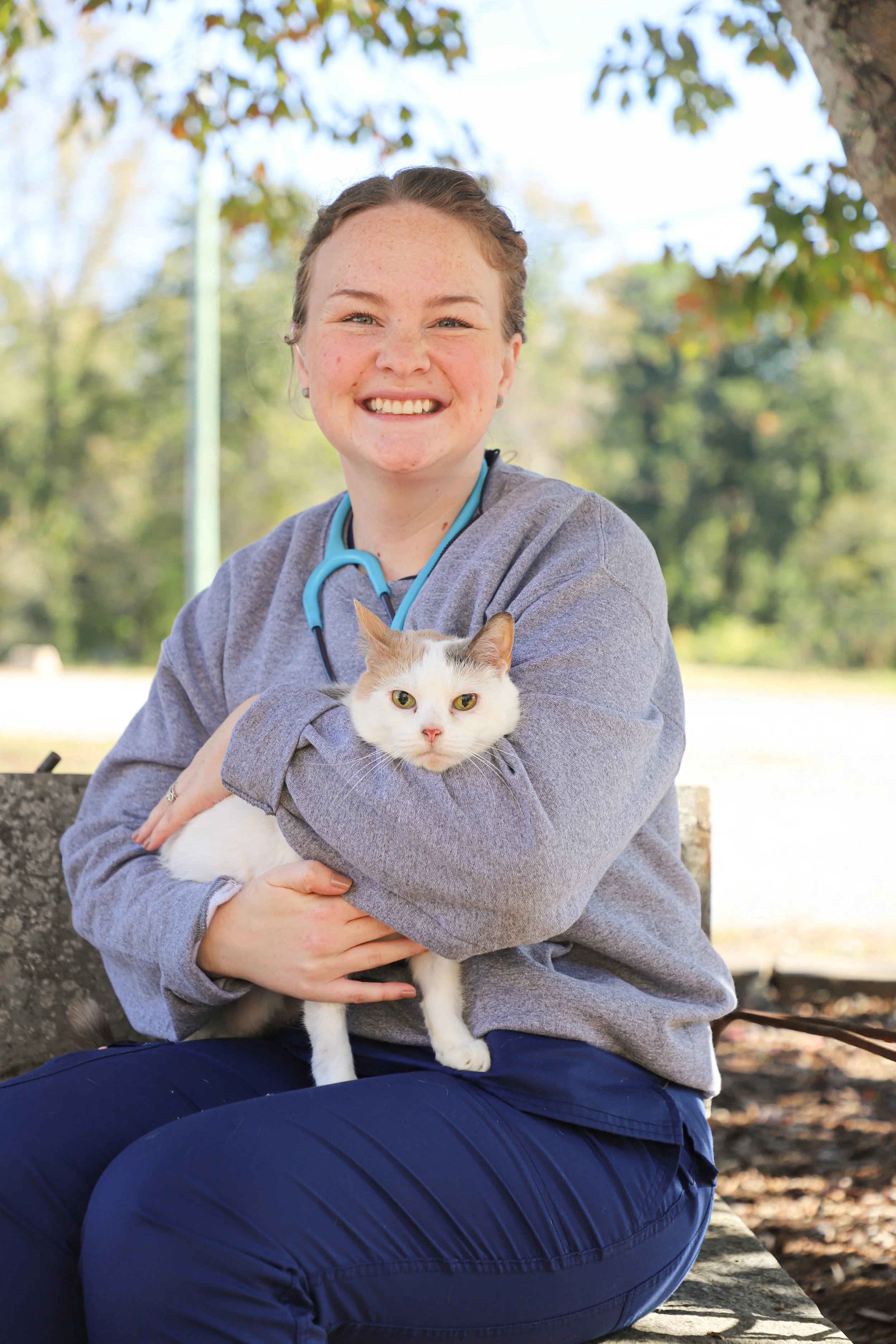 A smiling young woman in medical scrubs with a stethoscope around her neck sitting outdoors on a bench, holding a white cat with a grey patch on its head.