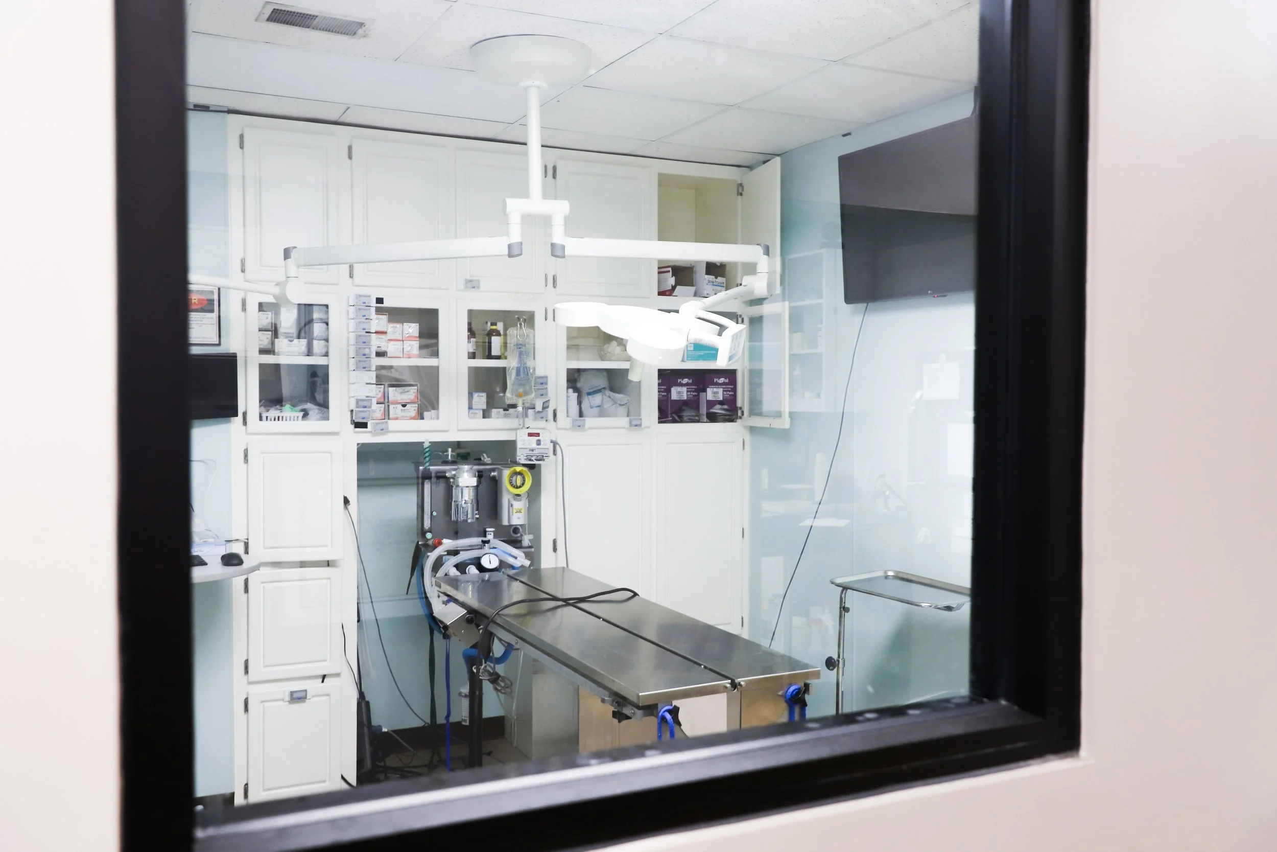 View through a window into a small medical operating room with a surgical table and medical equipment.