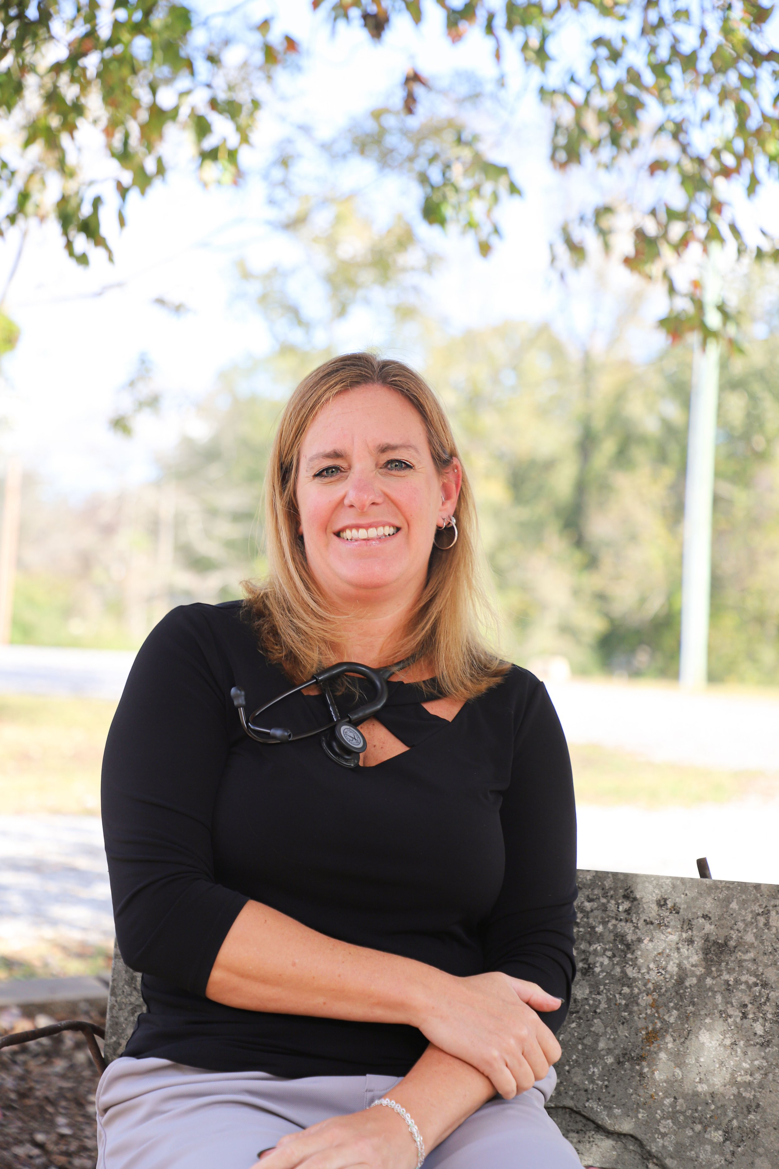 A woman with shoulder-length blonde hair, wearing a black top with cut-out details and gray pants, sitting on a stone bench outdoors, smiling at the camera under a tree with green leaves.