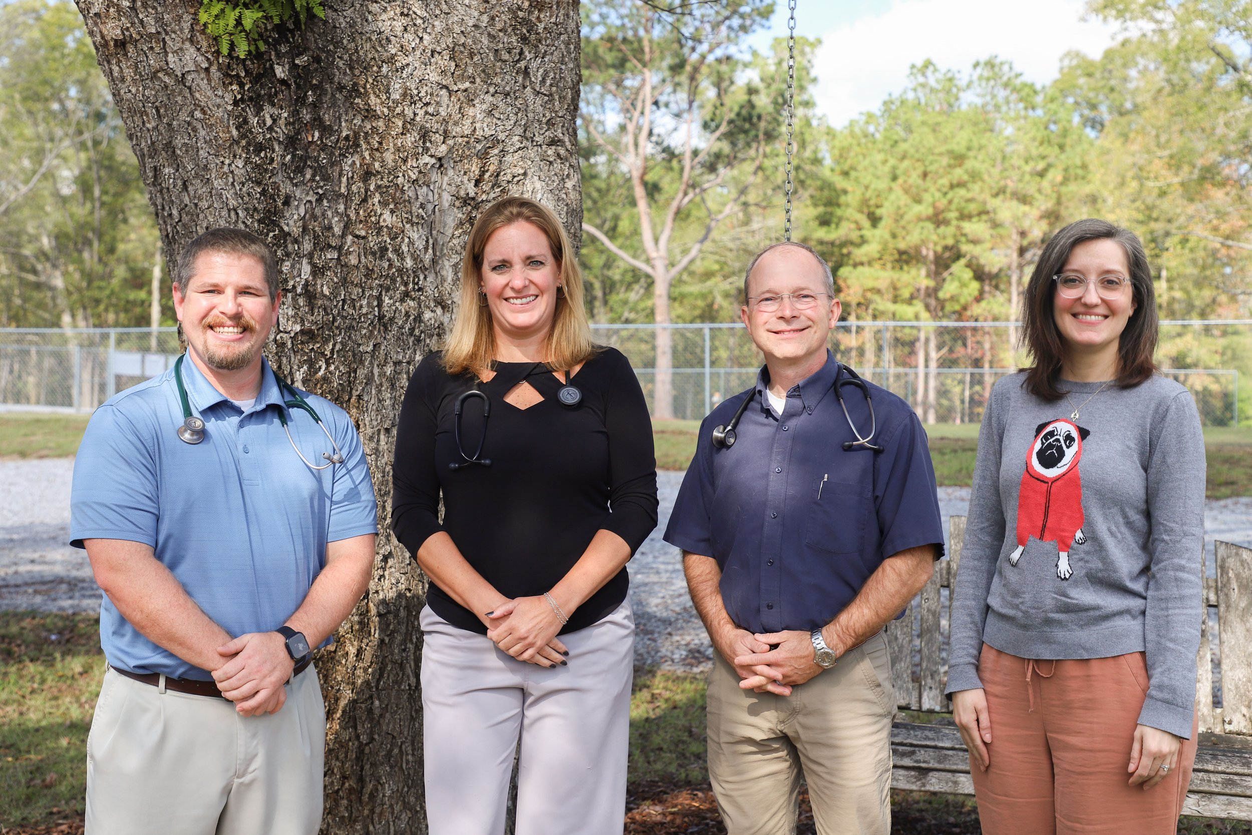 Four healthcare professionals standing outdoors in front of a large tree, smiling, with a chain-link fence and trees in the background.