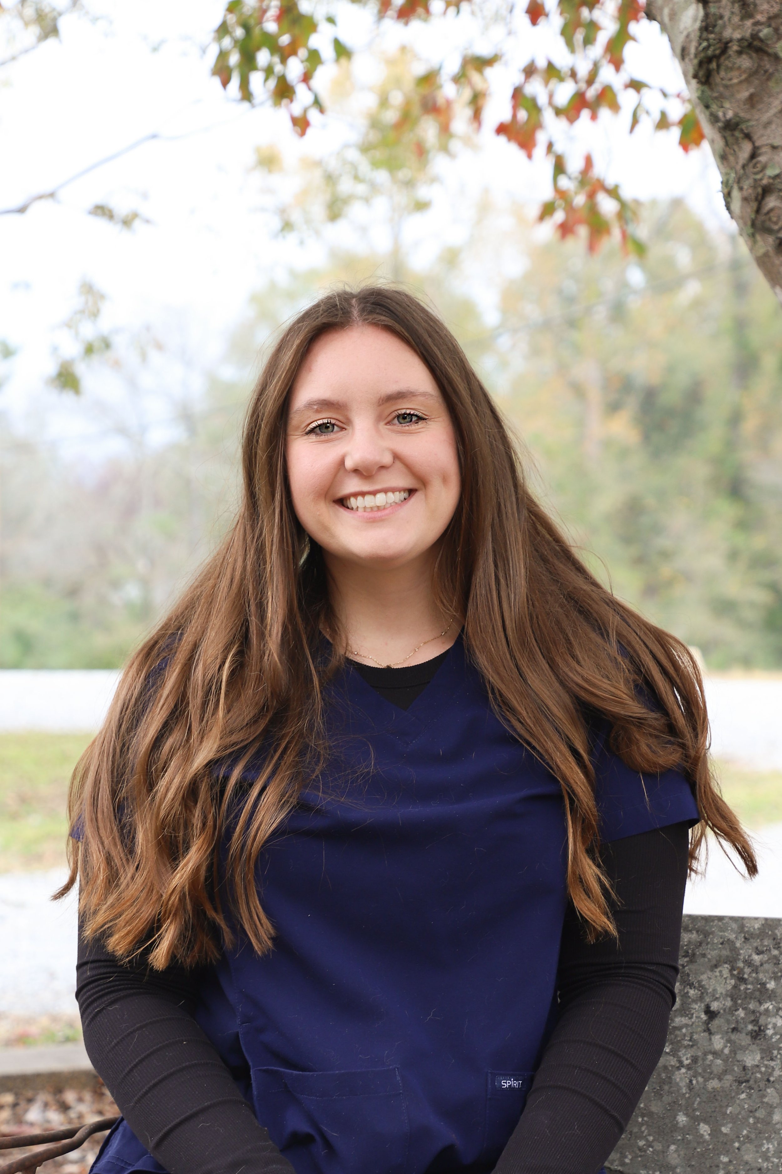 A young woman with long brown hair wearing a blue and black medical uniform smiling while sitting outdoors near a tree with fall foliage.