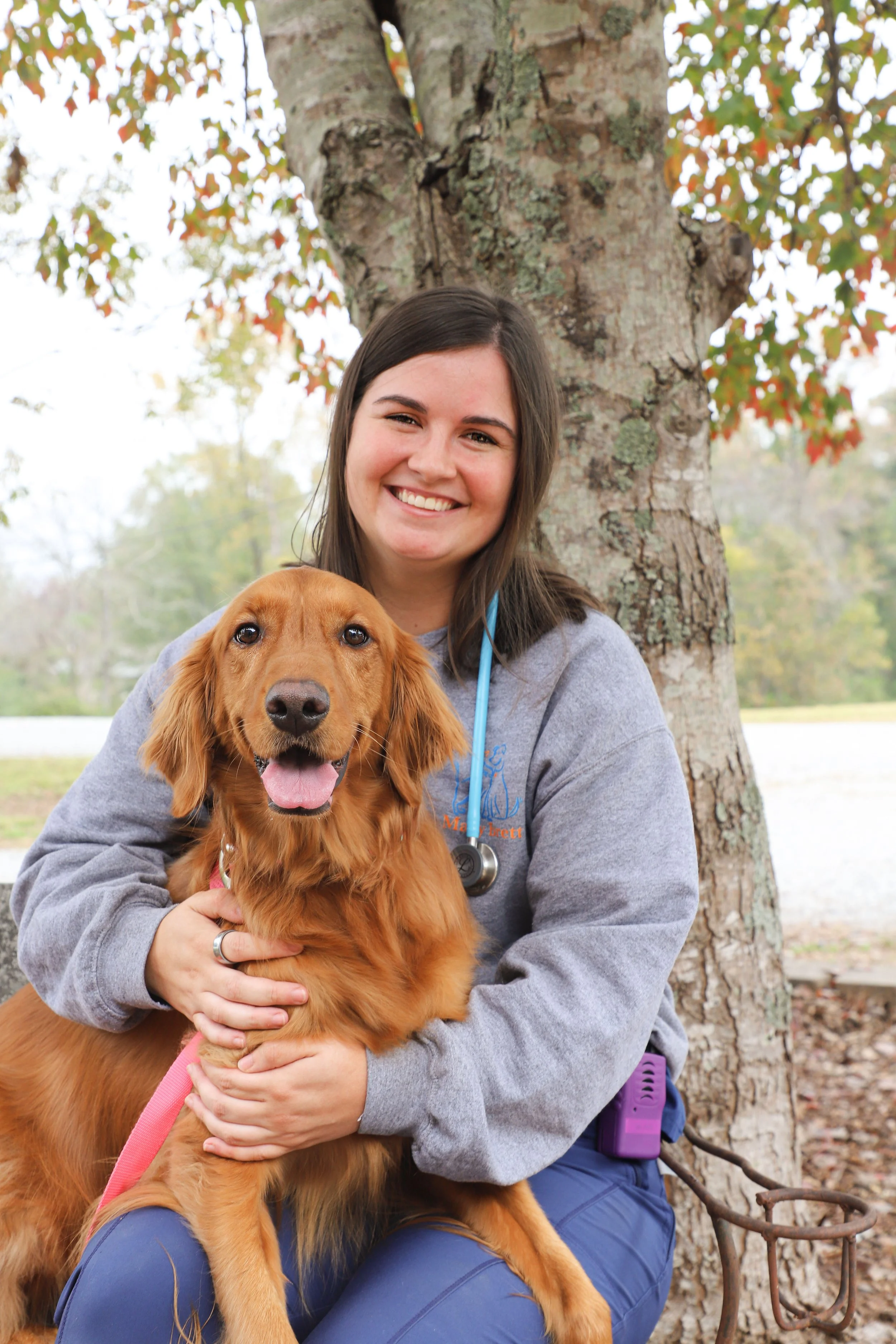 A smiling woman dressed as a veterinarian holding a golden retriever outdoors next to a tree in a park.