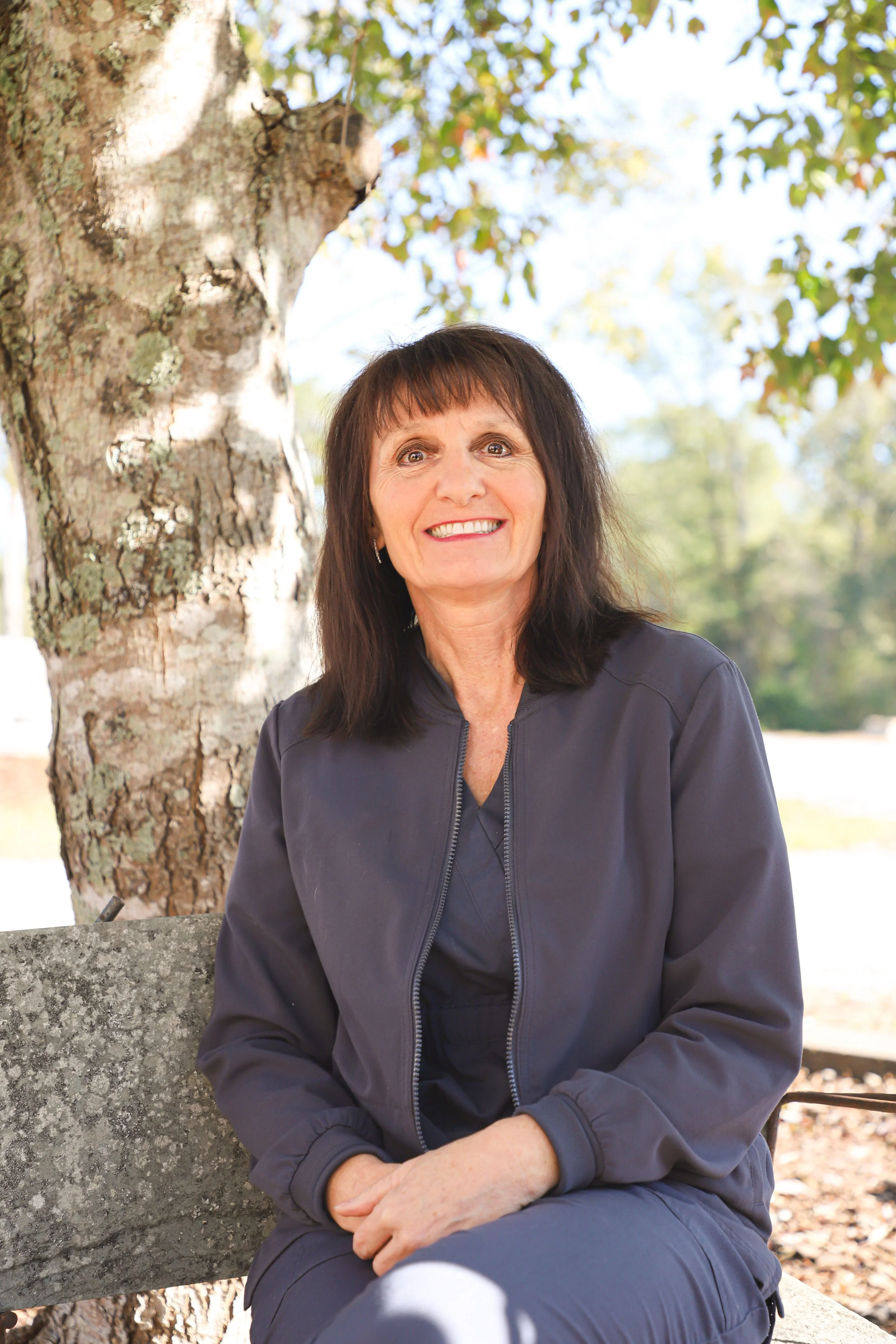 A smiling woman with dark hair, wearing a gray jacket, sitting on a bench outdoors with a large tree in the background.