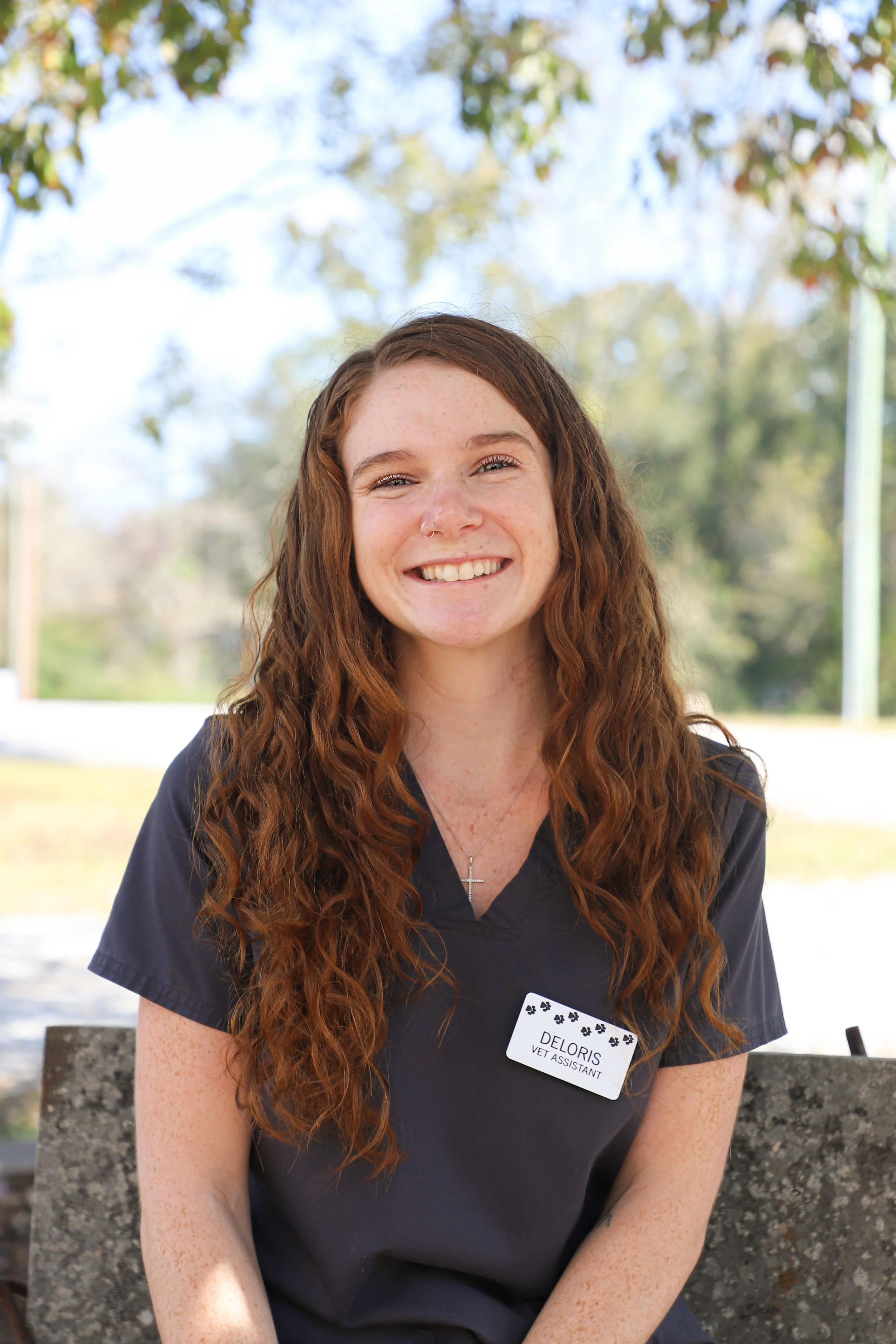 A young woman with long, curly red hair, smiling, wearing a dark uniform with a name tag that reads 'Deloris, Vet Assistant,' sitting outdoors on a concrete bench with trees and sky in the background.