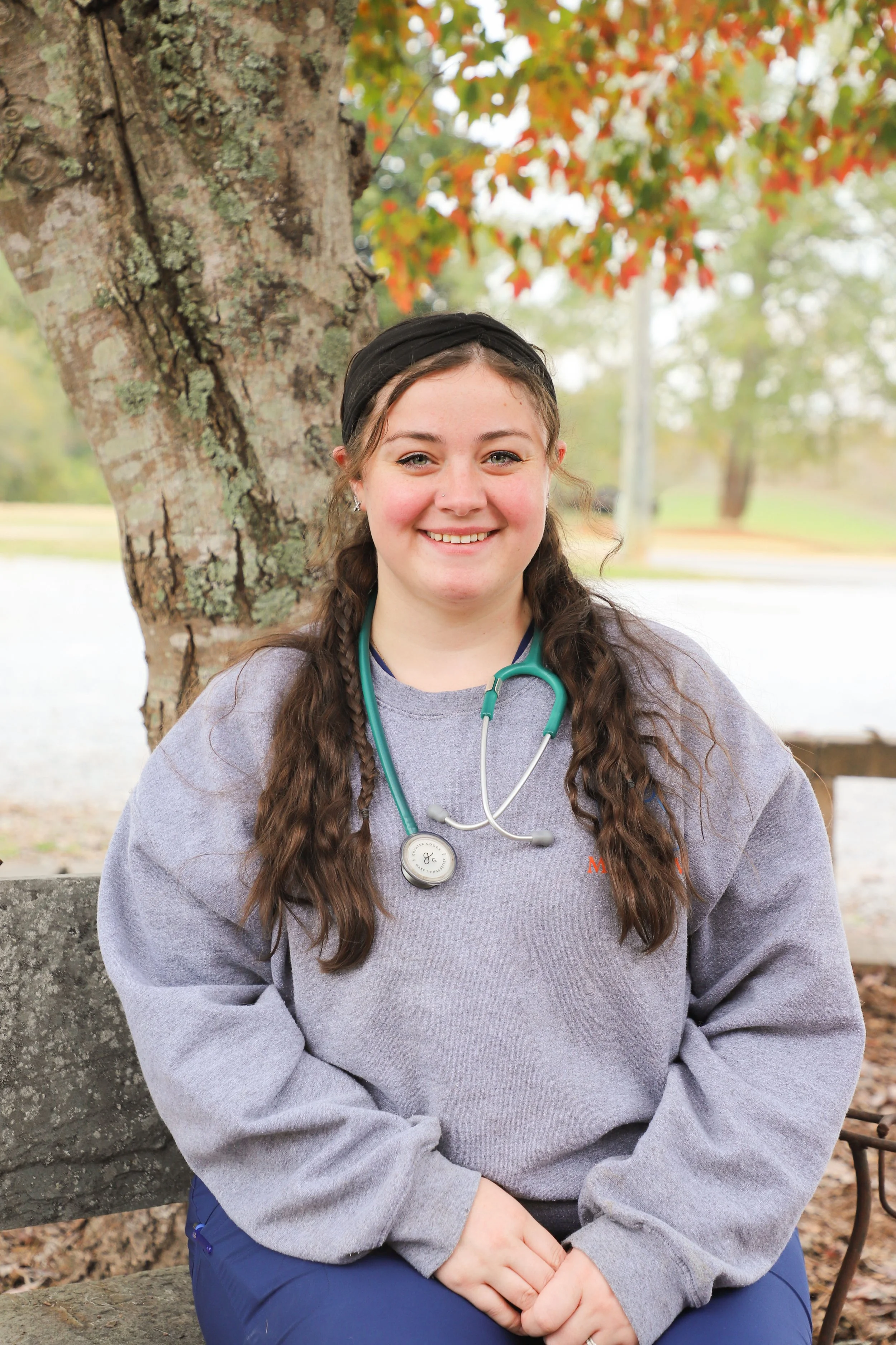 Young woman with long wavy brown hair, smiling, wearing a gray sweatshirt with a stethoscope around her neck, sitting outdoors in front of a tree with autumn leaves.