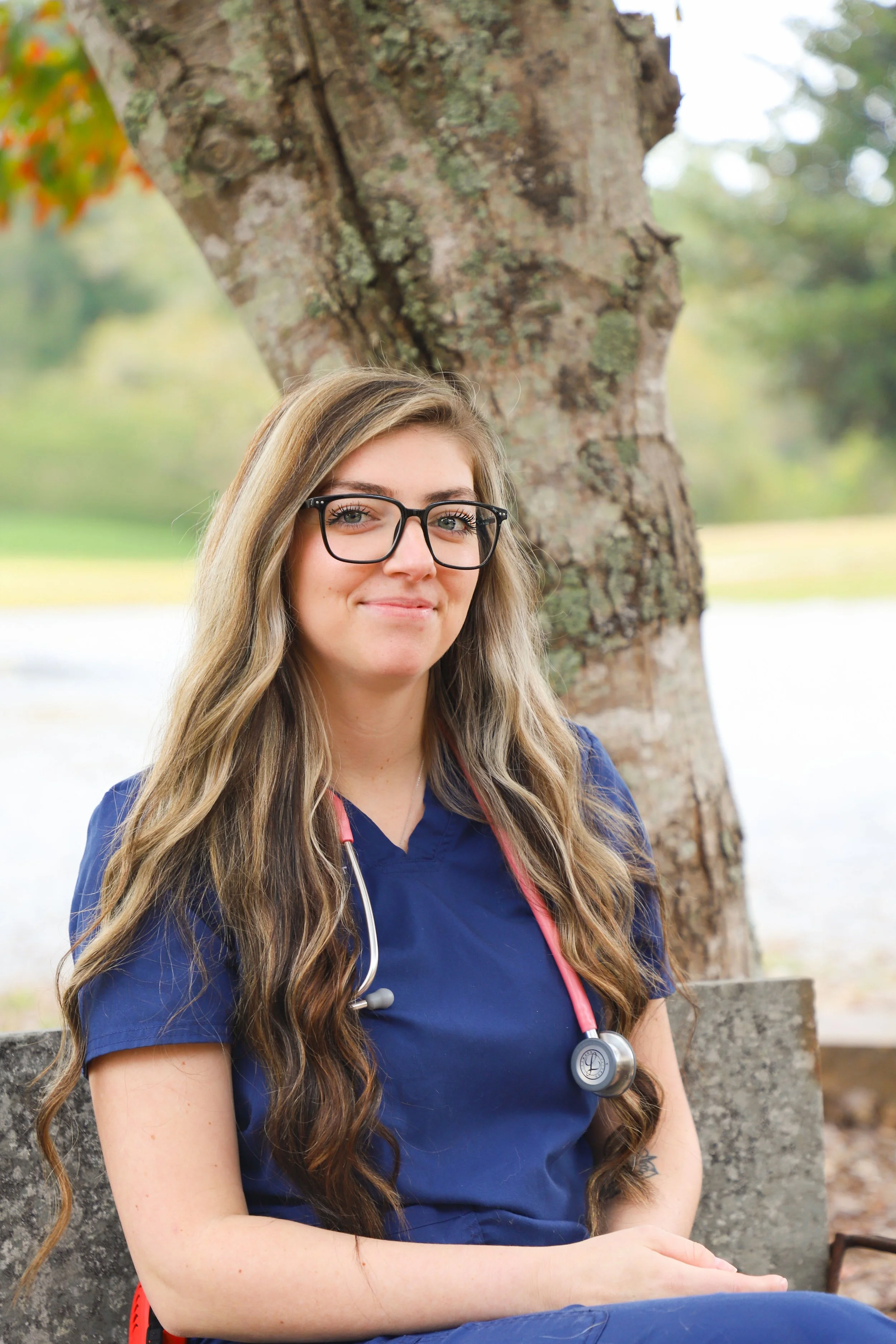 A young woman with long wavy hair wearing glasses and blue medical scrubs, sitting outdoors near a large tree with a stethoscope around her neck, smiling.