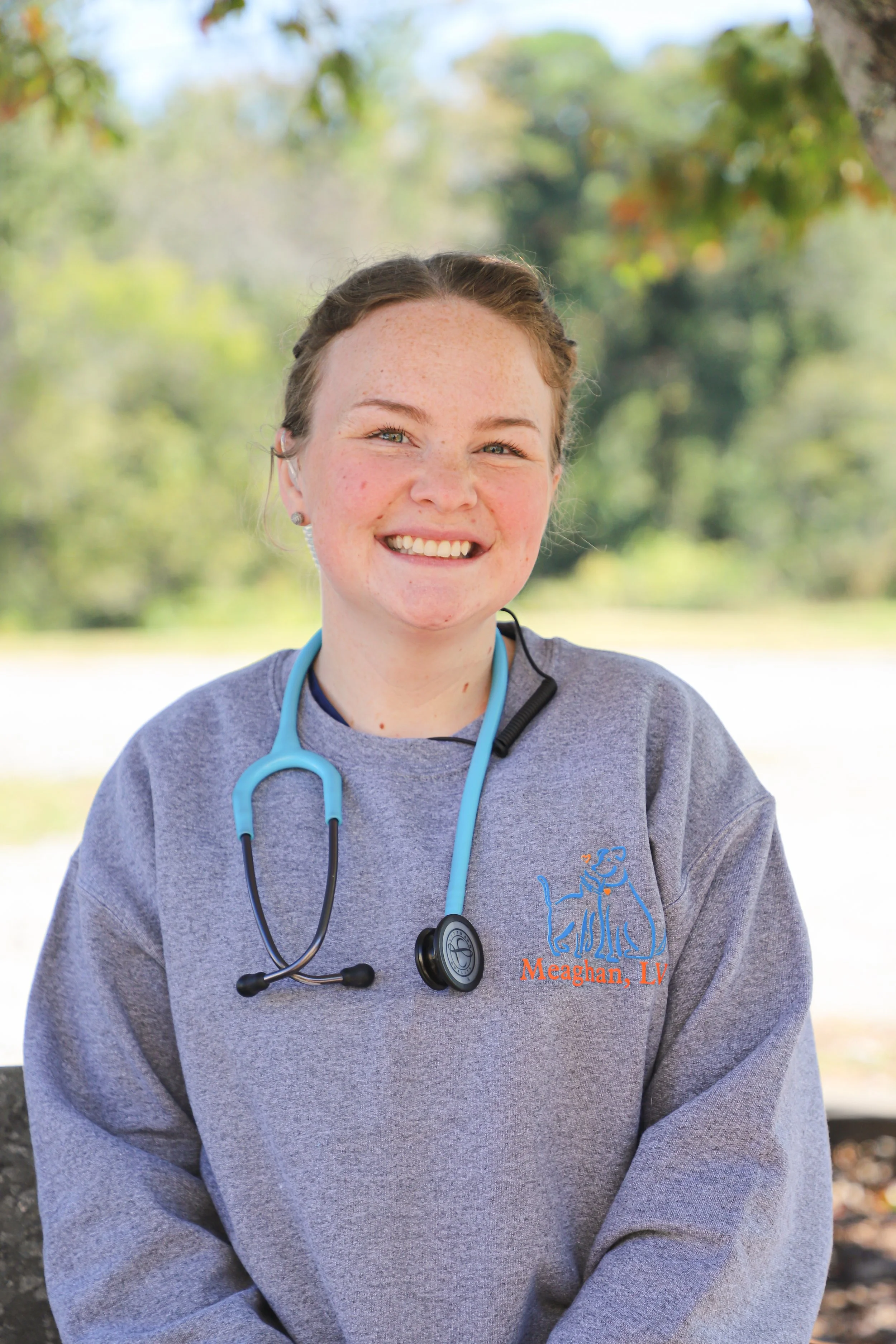 A smiling young woman wearing a gray sweatshirt with a veterinary logo, standing outdoors with trees in the background, and a stethoscope around her neck.
