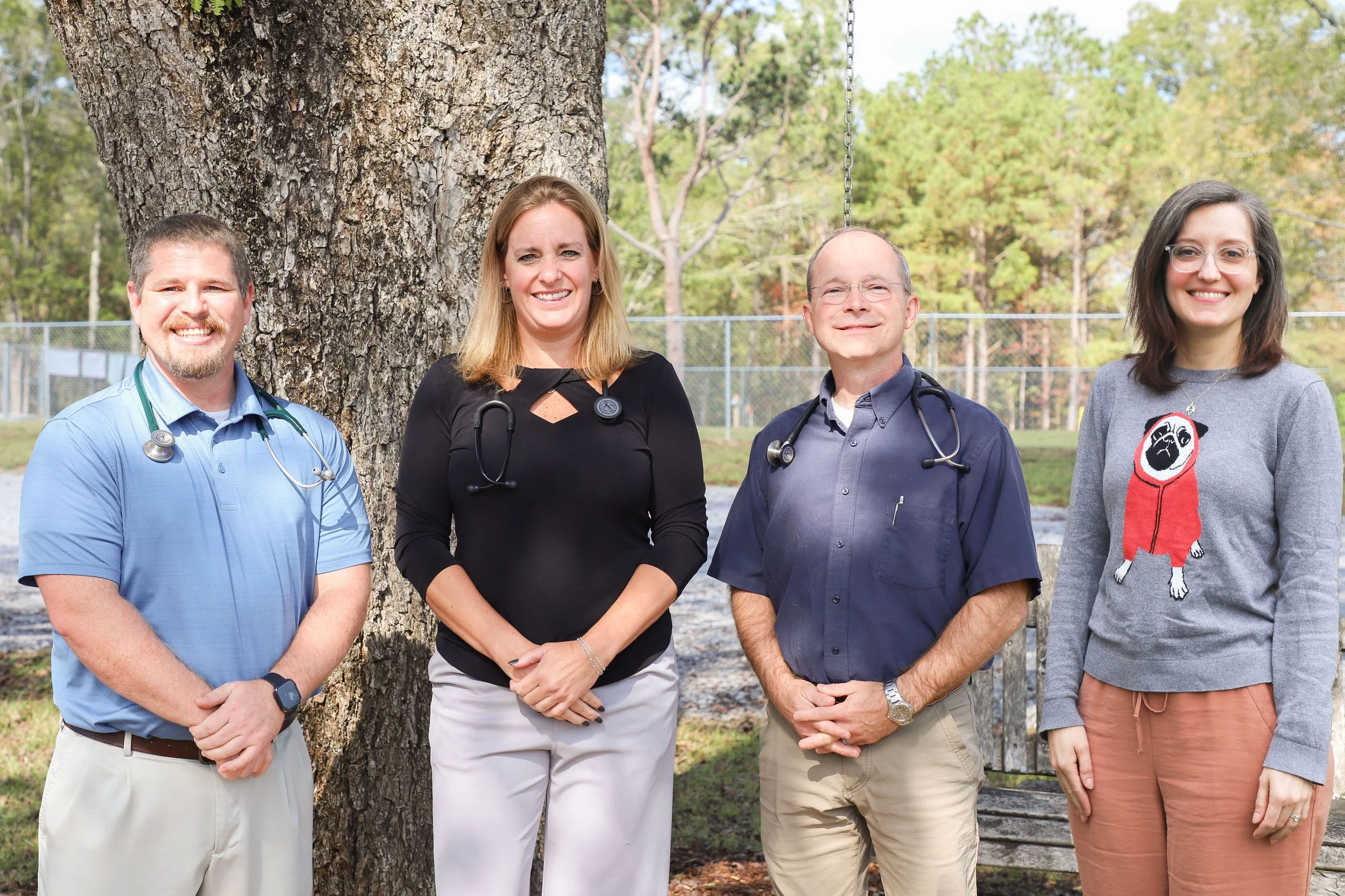 Four medical professionals standing outdoors in front of a large tree, smiling, with a playground swing hanging nearby.
