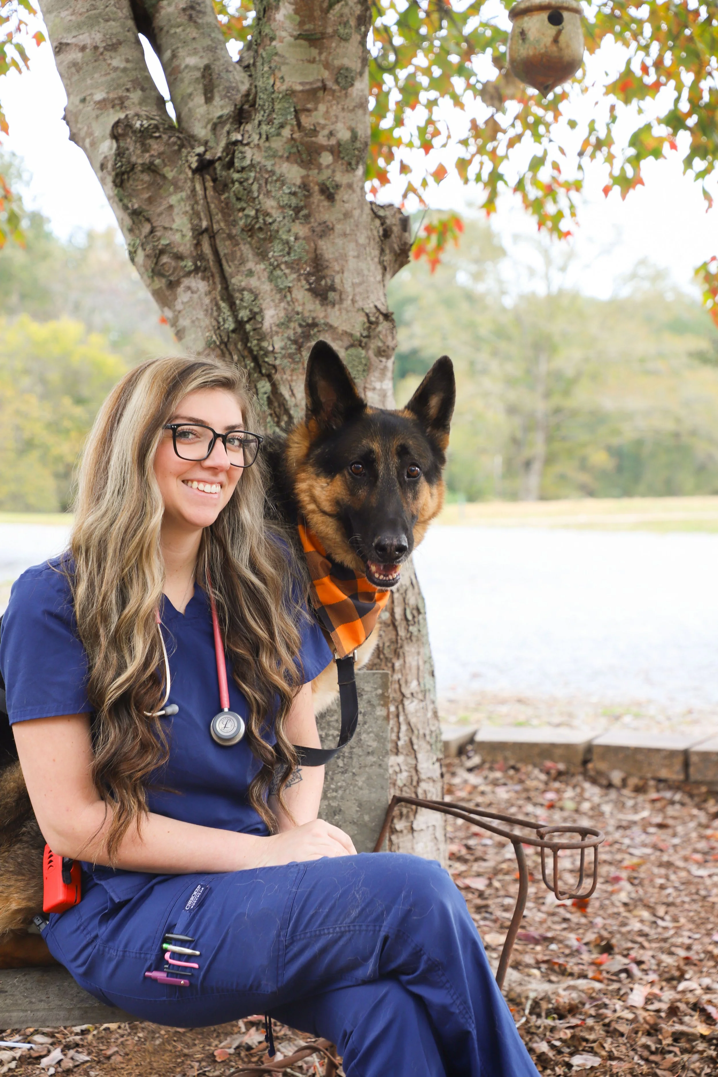 A young woman in medical scrubs and glasses sitting on a bench beside a large dog with black and tan fur, both smiling in an outdoor park setting with a tree, water, and autumn leaves in the background.