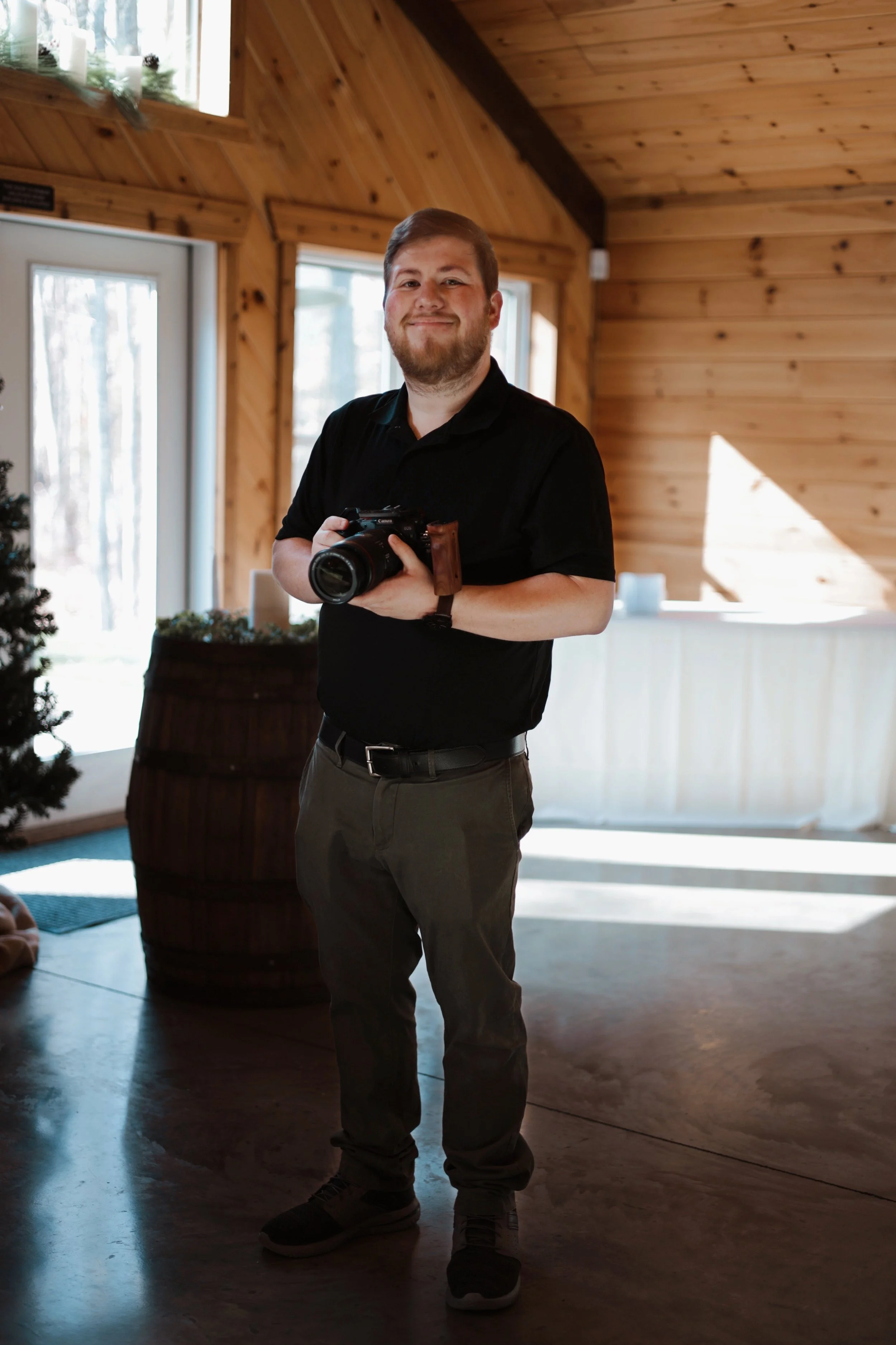 A man with a beard and short hair, wearing a black polo shirt, stands holding a camera in his hands and smiling.