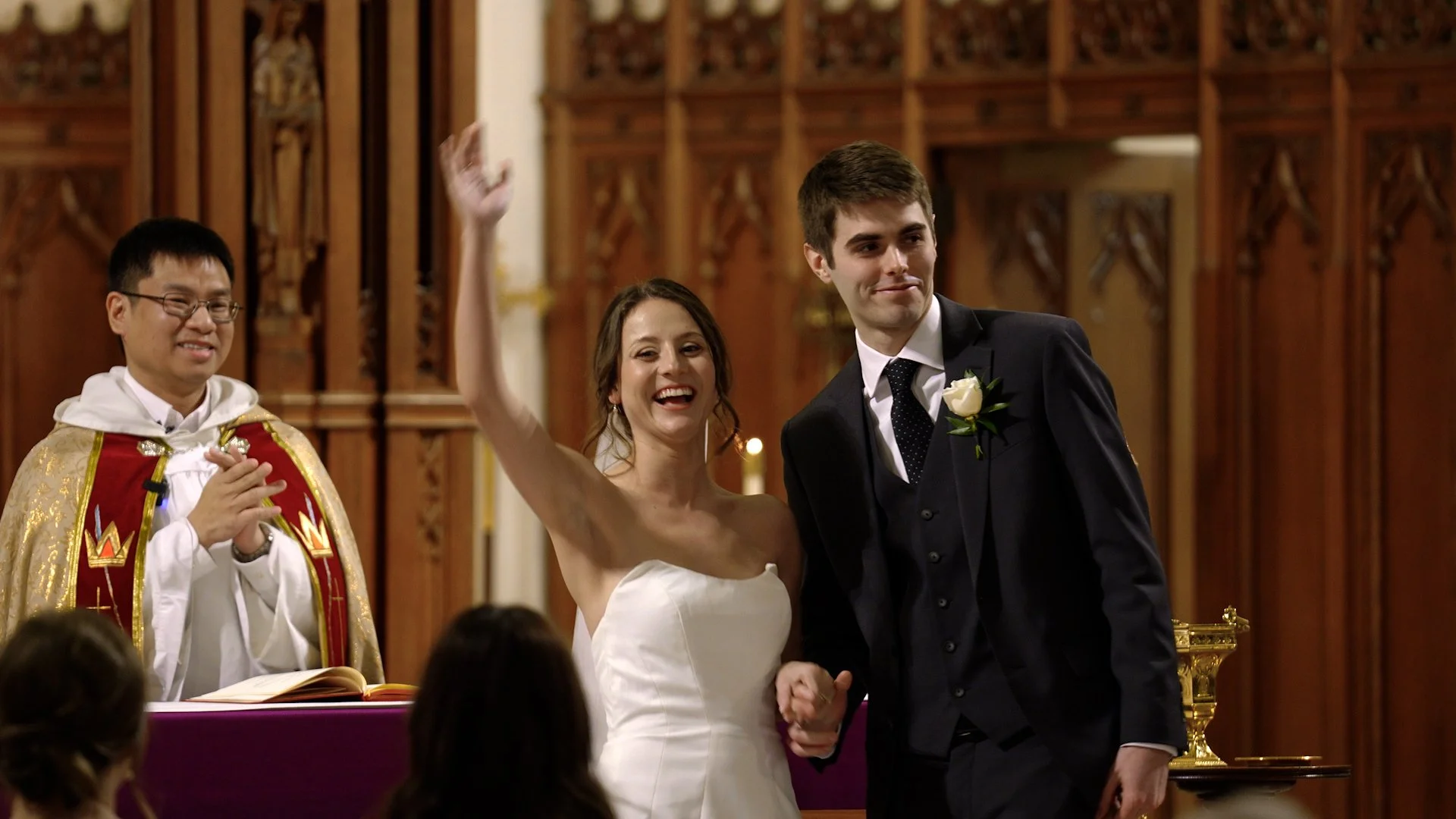 A bride and groom smiling and holding hands during their wedding ceremony in a church, with a priest standing nearby and people in the audience watching.