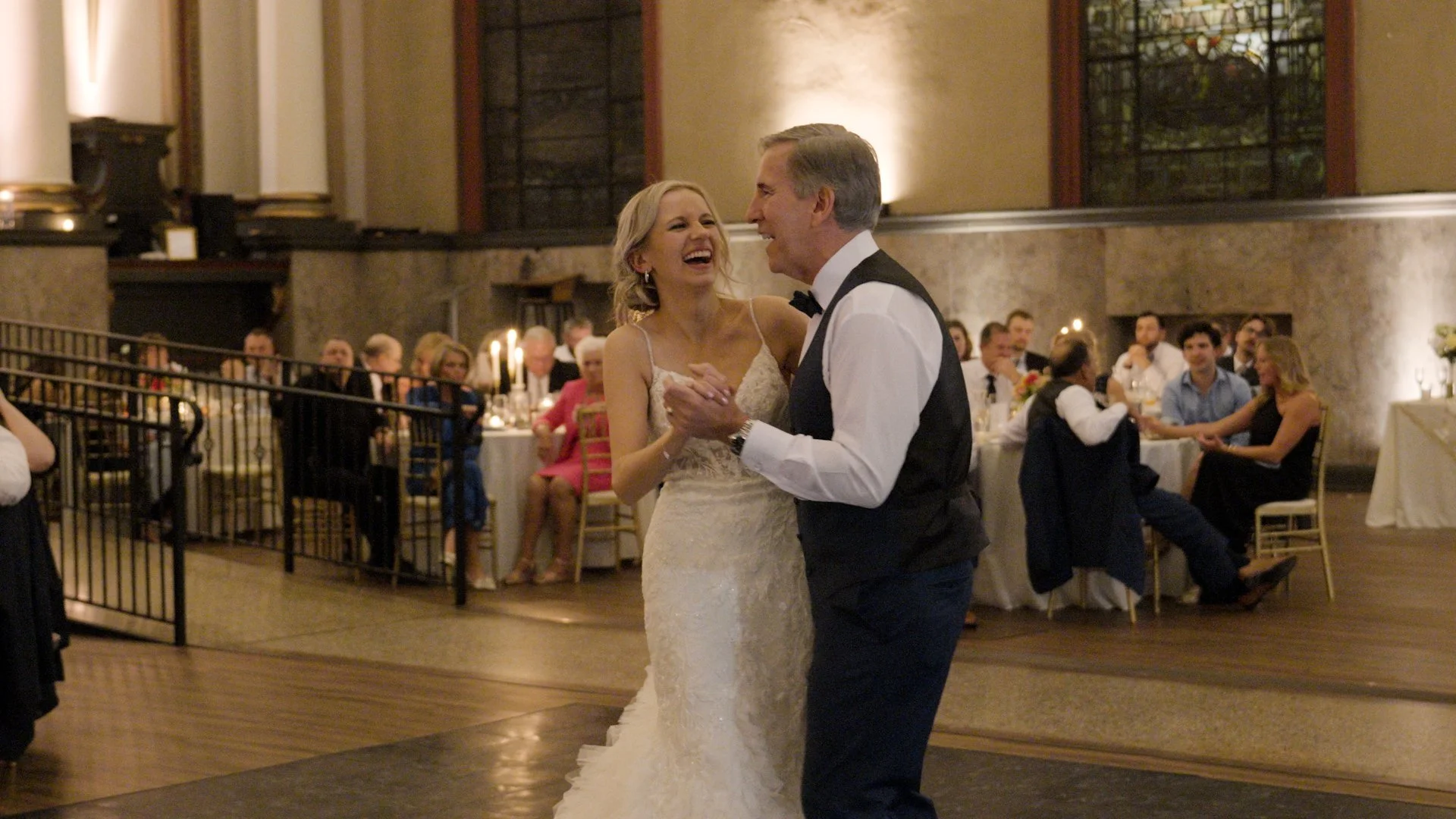 A bride and groom dance at their wedding reception, smiling and laughing in an elegant hall with seated guests in the background.