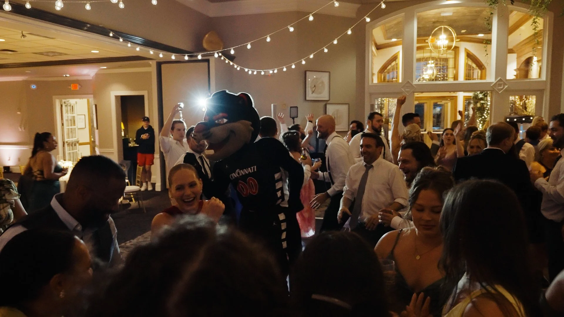 People dancing and celebrating at a lively indoor party with string lights and large windows showcasing the interior decor.
