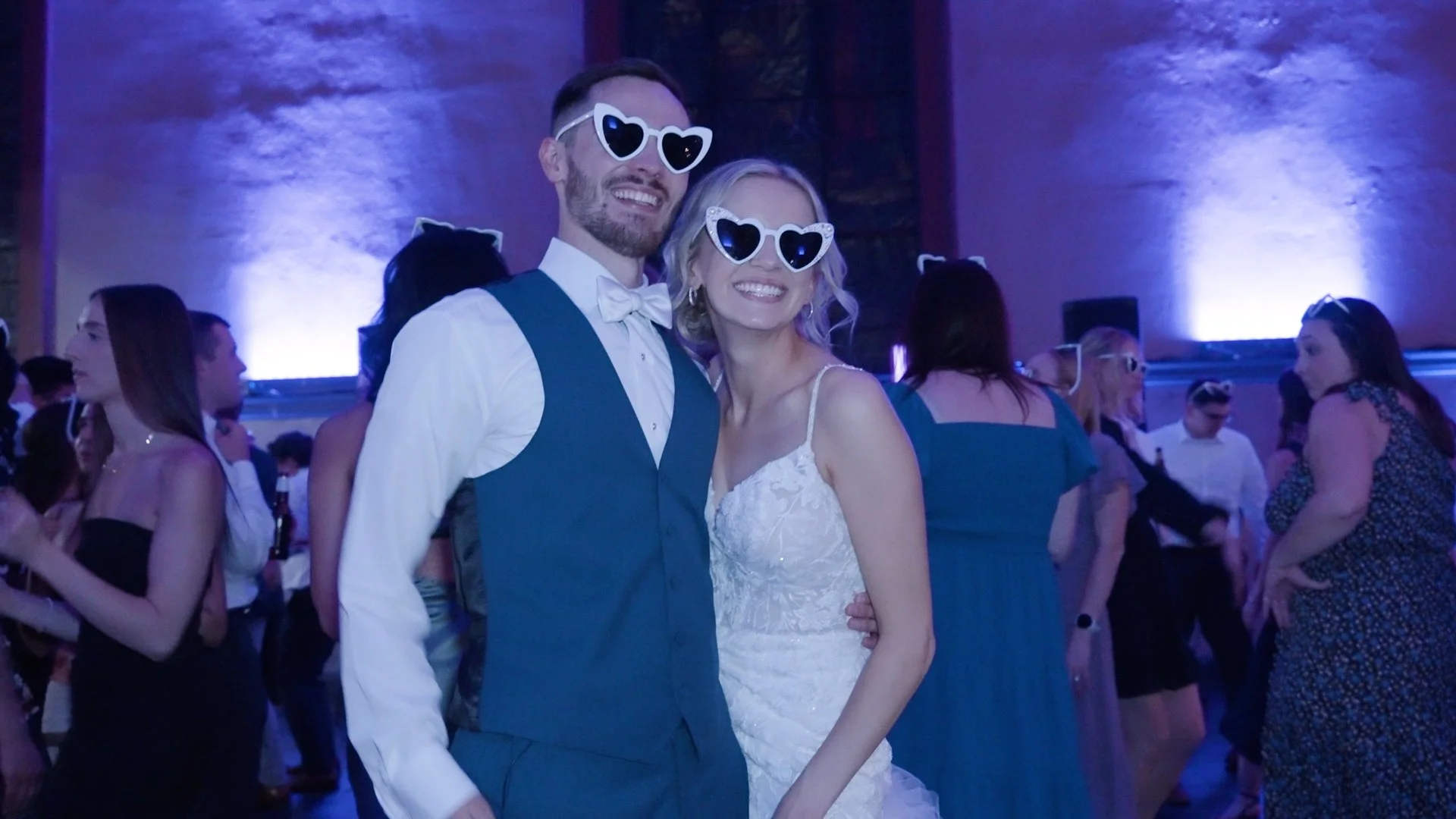 A wedding reception with a smiling bride and groom wearing heart-shaped sunglasses, surrounded by guests in a dimly lit venue.
