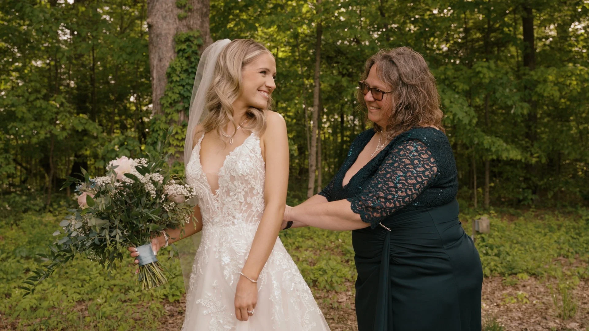A bride in a lace wedding gown holding a bouquet of flowers, smiling as her mother helps her into her dress, outdoors in a wooded area.