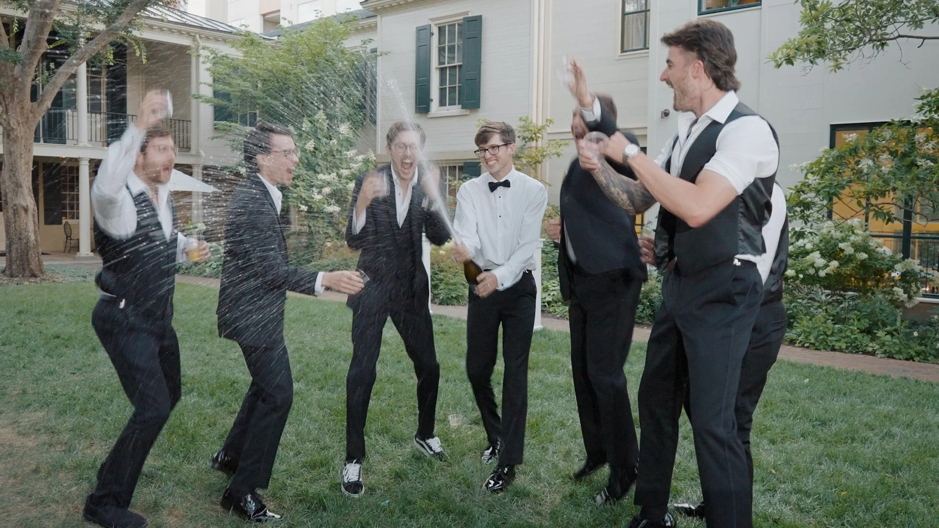 Group of friends dressed in formal attire celebrating outdoors, spraying champagne and laughing in a garden area with trees and a white house in the background.