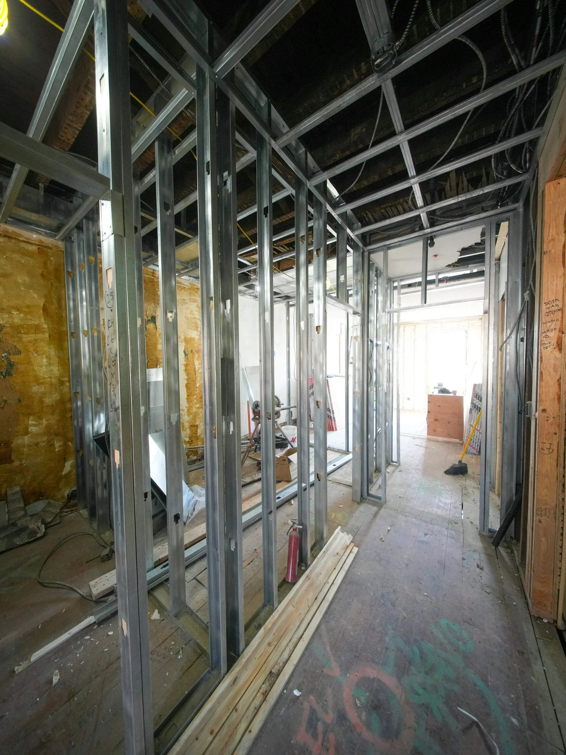 Interior view of a building under construction with metal framing, exposed wiring, and construction tools and materials on the floor.