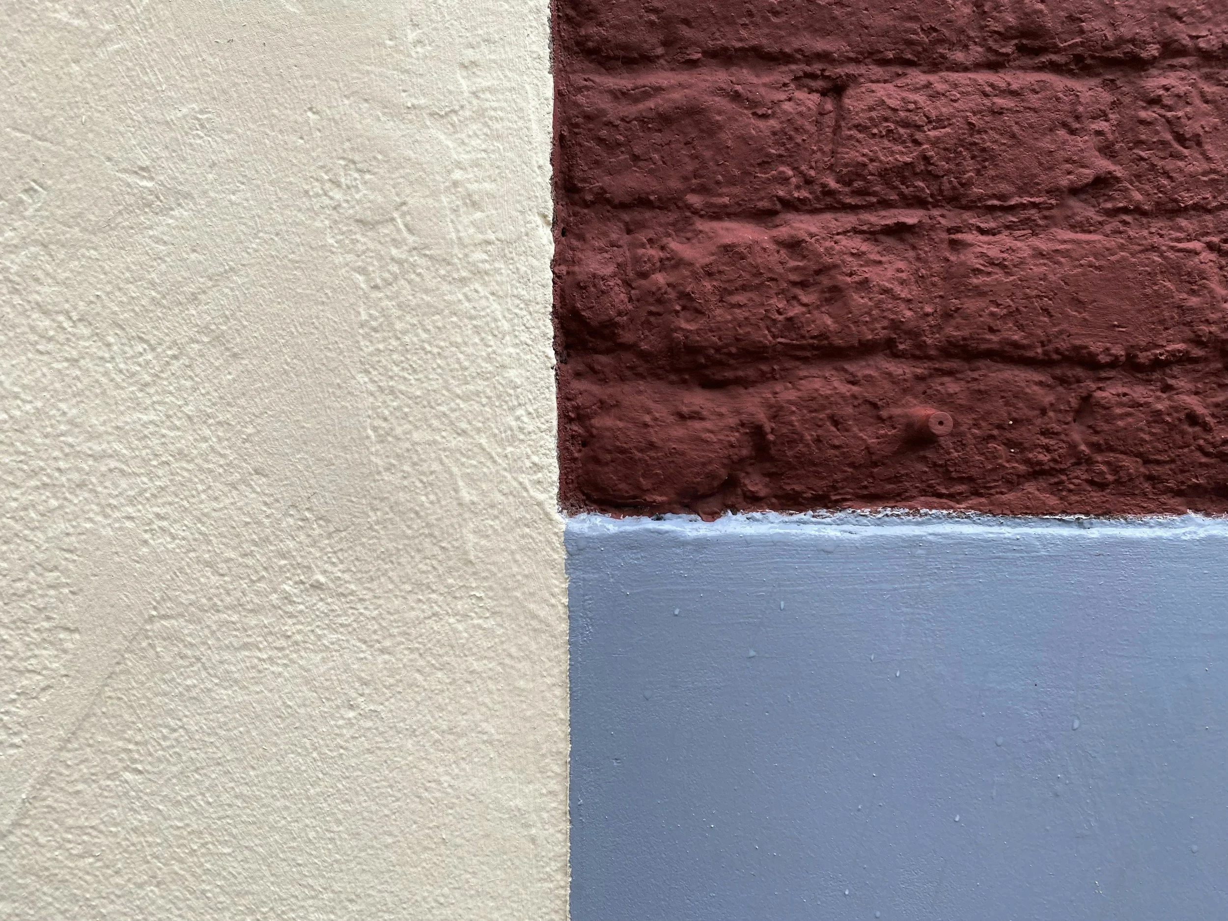 Close-up of a wall with beige textured paint on the left side, and a red brick wall with a rough texture on the right side. There is a thin horizontal strip of blue paint at the bottom of the red brick wall.