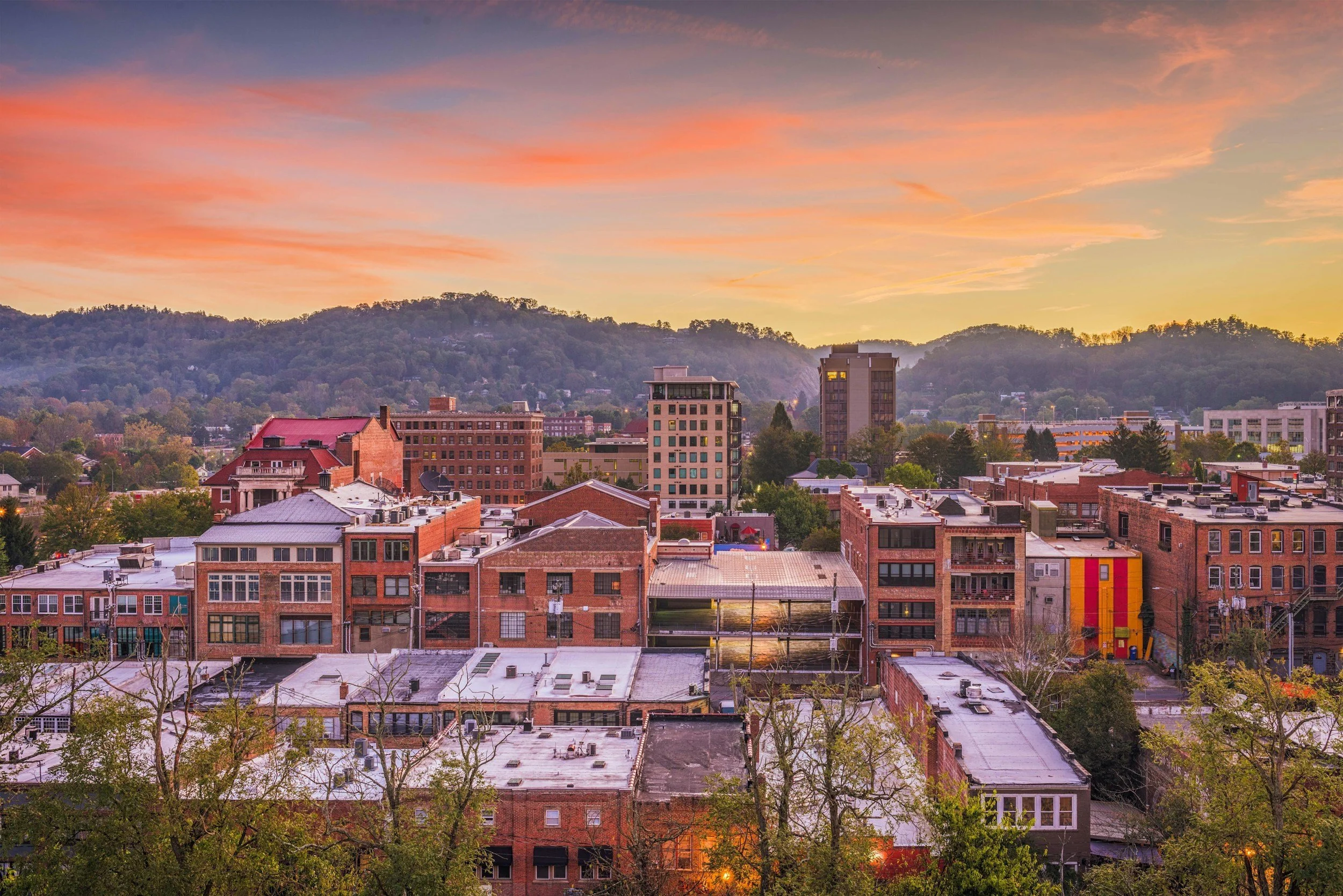 A cityscape at sunset showing various buildings, trees, and rolling hills in the background with a colorful sky.