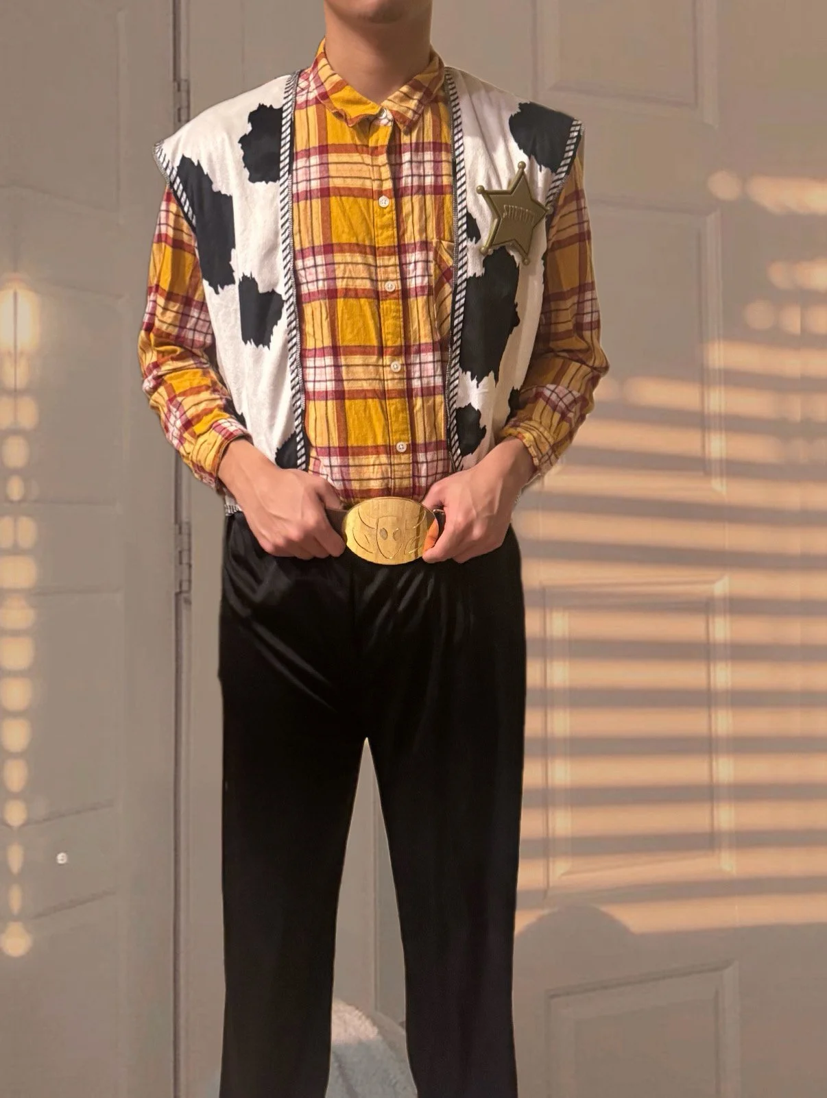 Person dressed as a sheriff with a cow-print vest, plaid shirt, sheriff badge, and a large belt buckle, standing indoors with sunlight casting shadows.