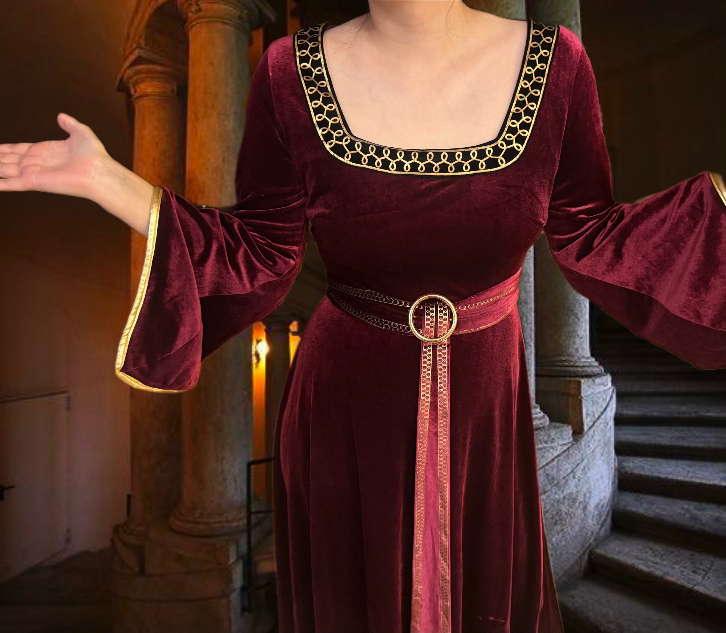 A person wearing an elegant red velvet dress with gold embroidery details on the neckline, open arms, and a matching belt with a gold buckle, standing in a historic setting with stone pillars and stairs.