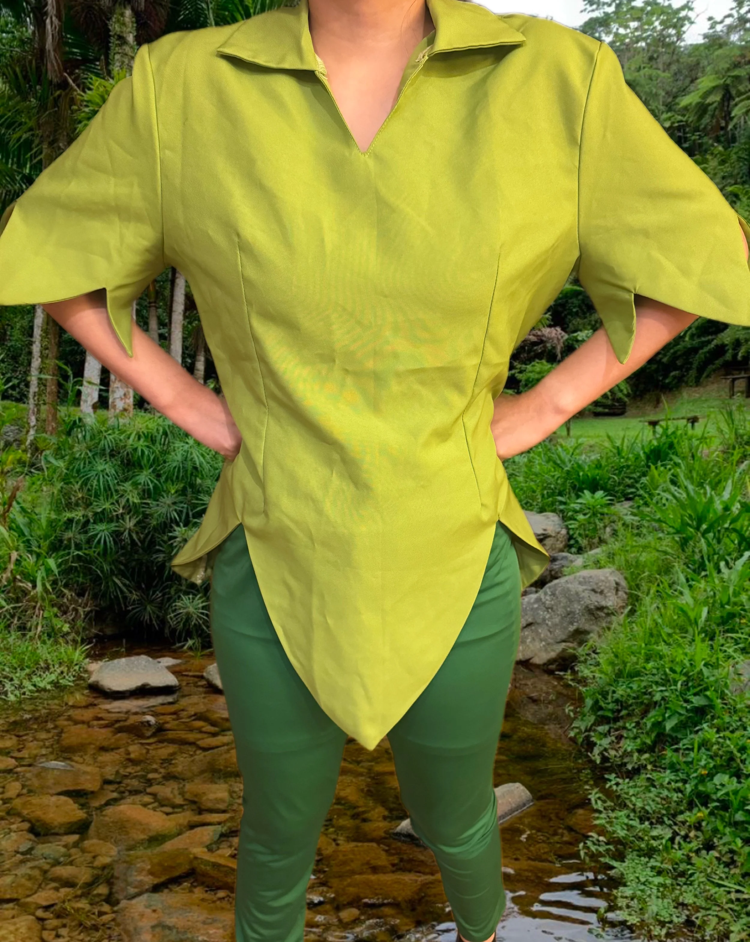 A person wearing a lime green shirt with puffed sleeves and a pointed hem, standing outdoors in a lush, green garden with rocks and plants in the background.