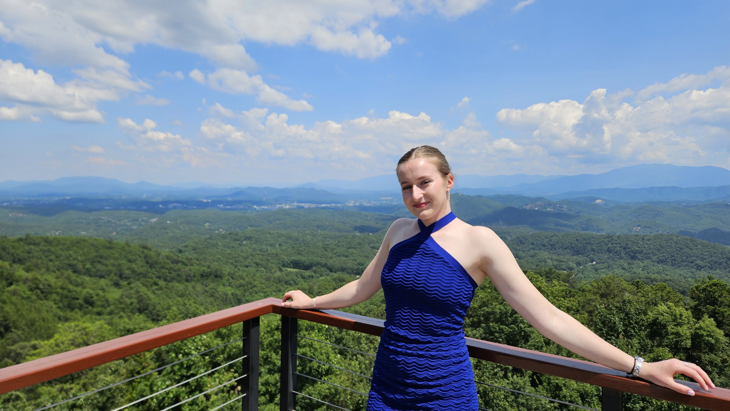 A young woman in a blue dress standing on a balcony overlooking a green landscape with rolling hills and mountains in the distance under a blue sky with fluffy white clouds.