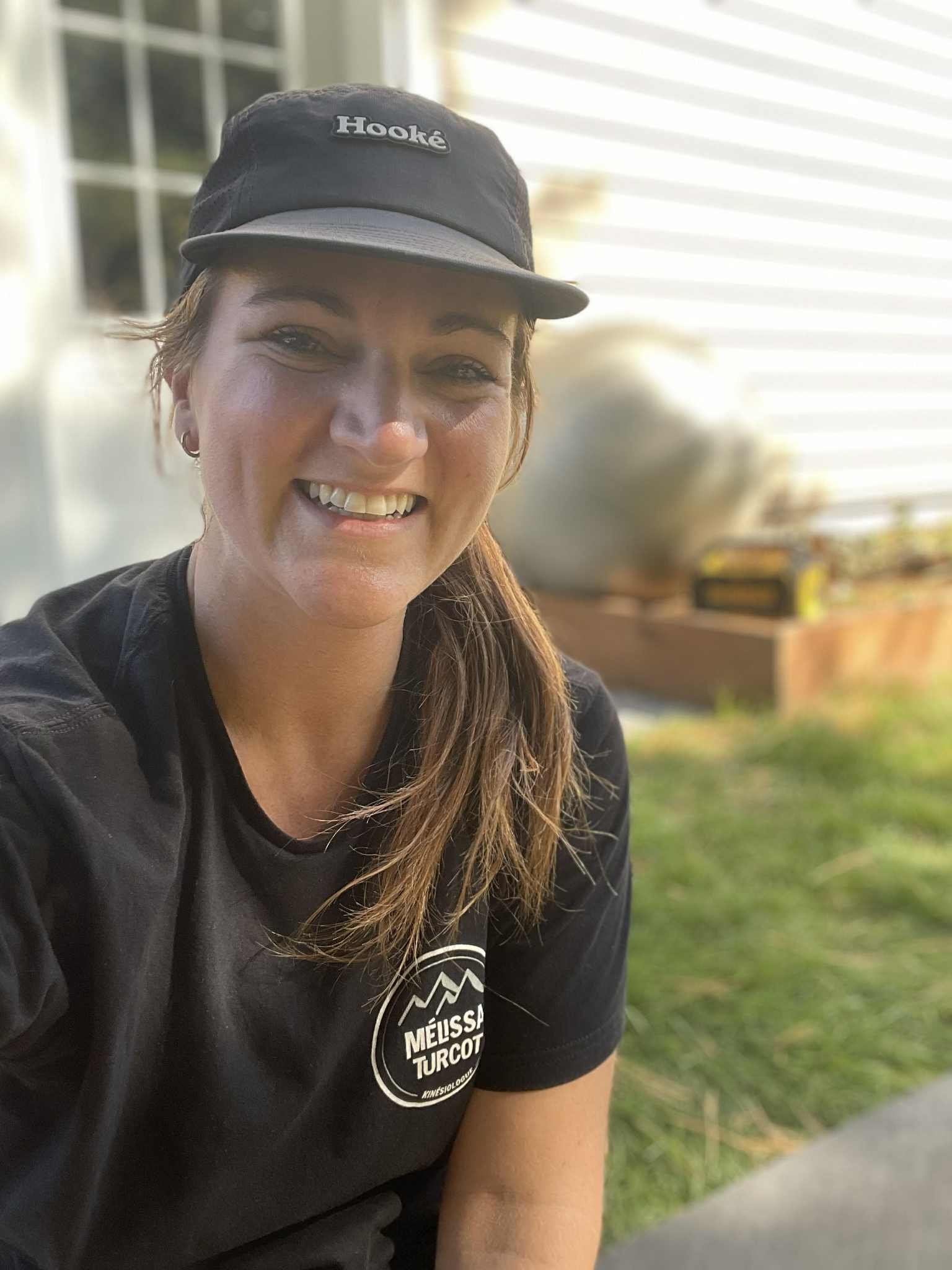 Une femme souriante portant une casquette noire avec le mot 'Hooké' et un t-shirt noir avec le logo 'MÉLISSA TURCOT' pose à l'extérieur, avec un fond flou d'une maison et de la végétation.