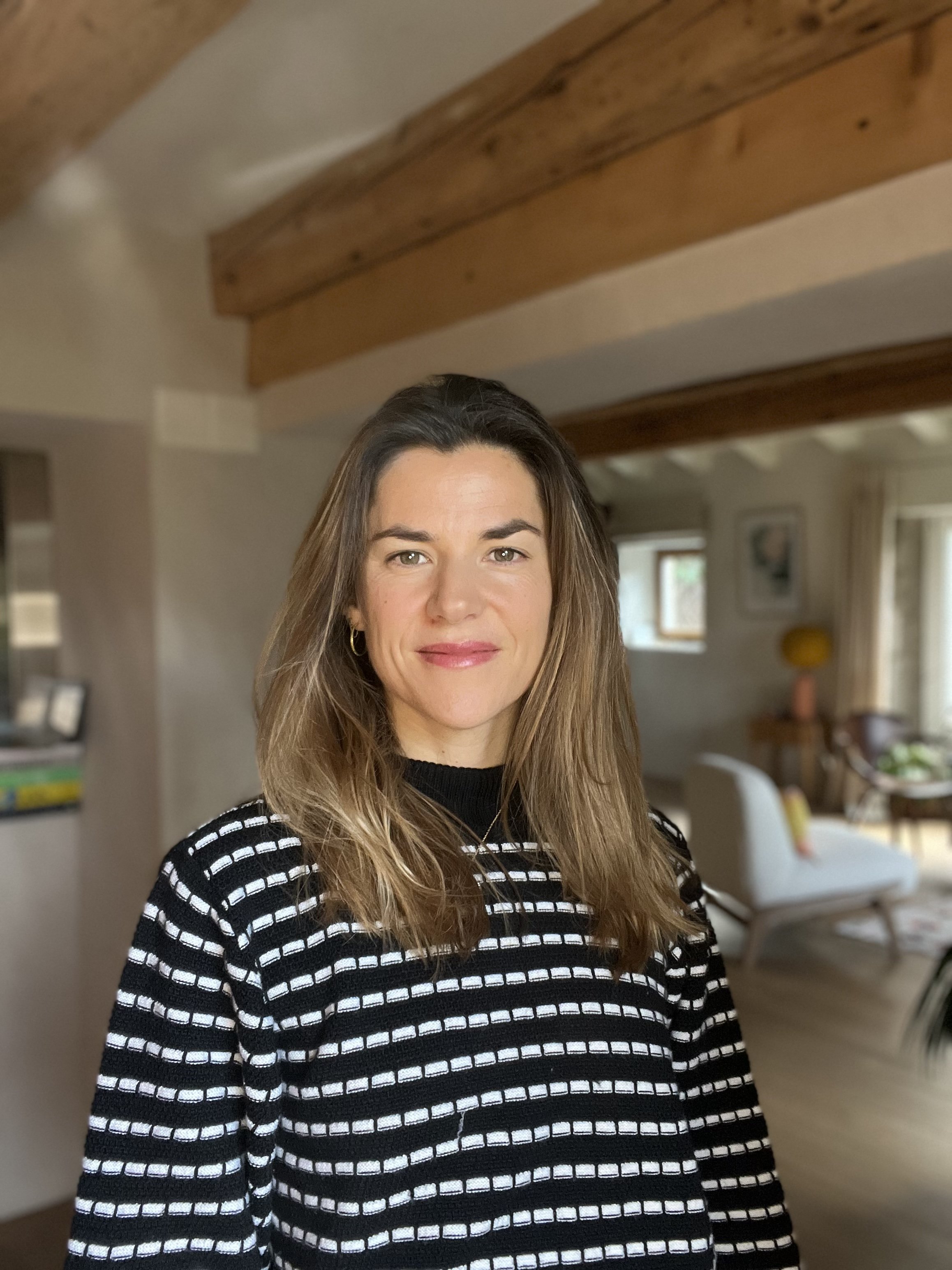 Une femme souriante, portant un pull à rayures noirs et blancs, dans un intérieur chaleureux avec des poutres en bois au plafond et un salon en arrière-plan.