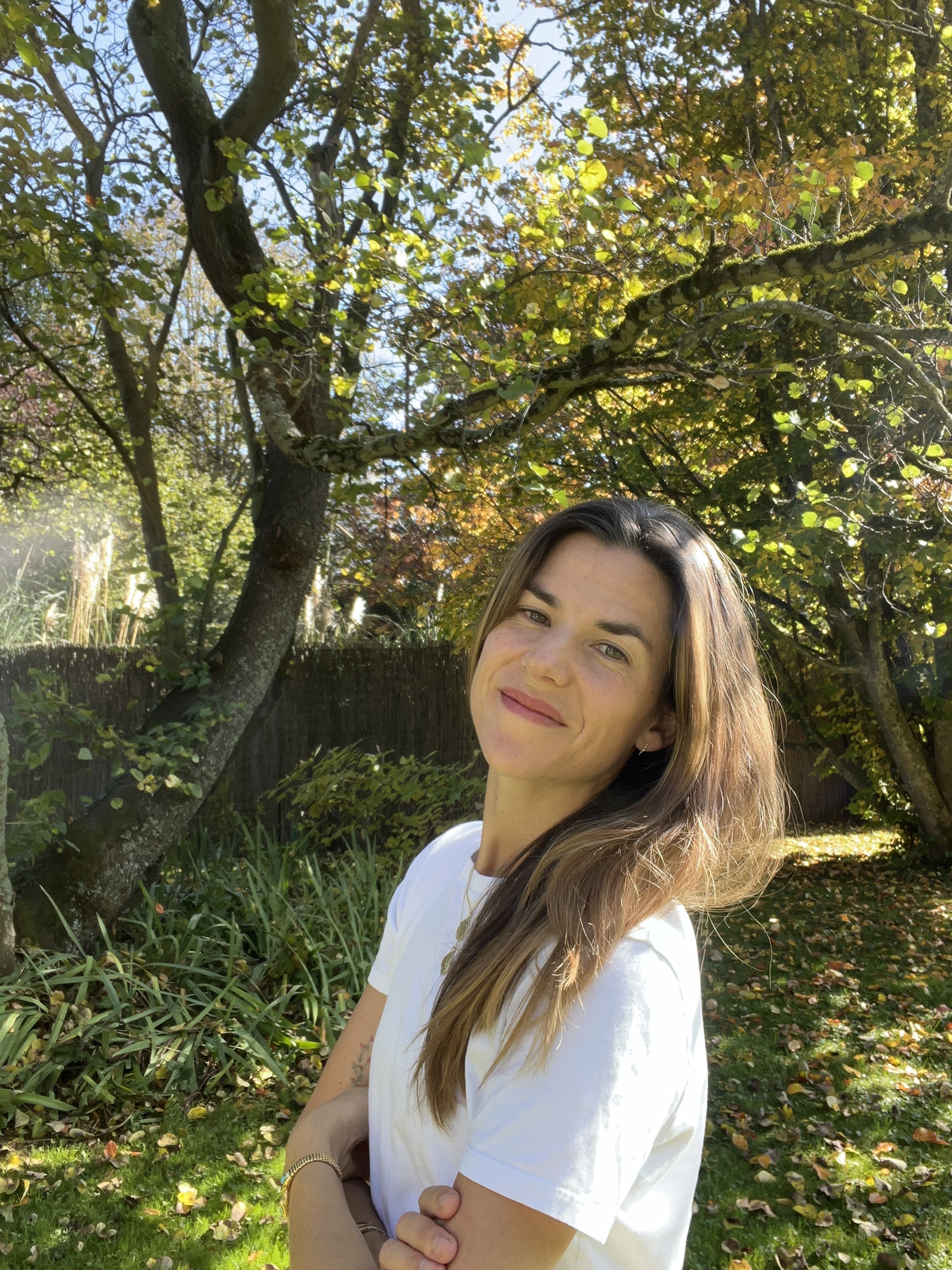 Une femme avec des cheveux longs et bruns, portant un t-shirt blanc, se trouve dans un jardin entouré d'arbres aux feuilles colorées en automne, sous un ciel ensoleillé.
