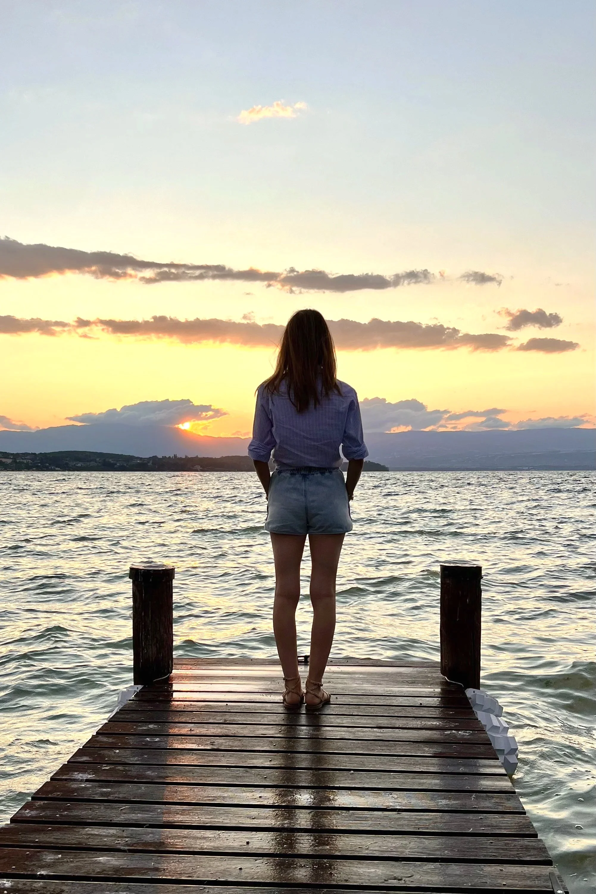 Une femme debout sur un quai en bois, regardant un coucher de soleil sur un lac.