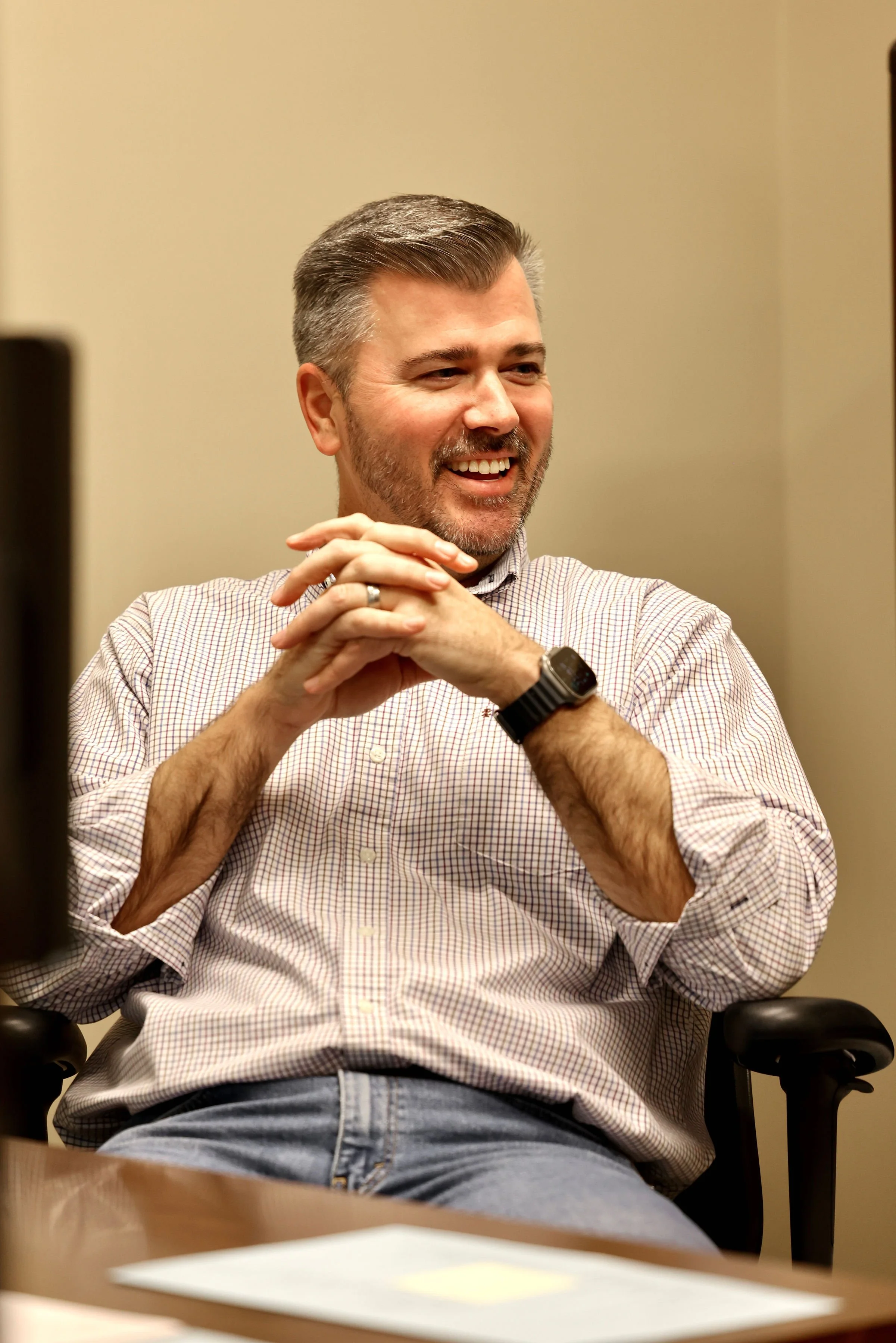 A middle-aged man with short gray hair and a beard, wearing a checkered shirt and a smartwatch, sitting at a conference table, smiling and leaning back with his hands clasped.