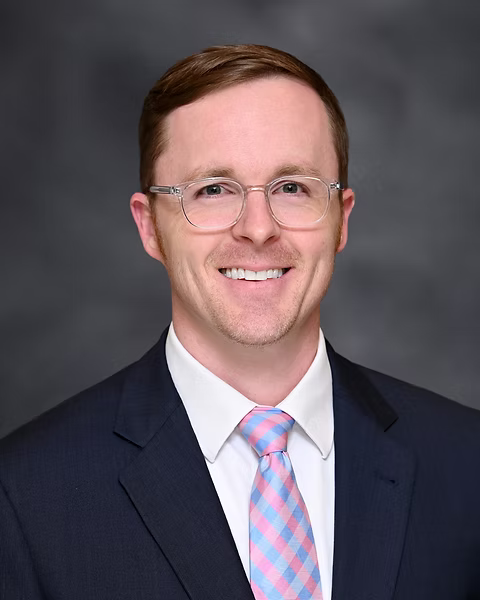 Professional headshot of a man with short brown hair, wearing glasses, a dark suit, white shirt, and pink patterned tie, smiling against a gray background.