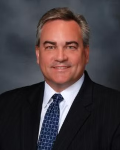 Professional headshot of a man with gray hair, wearing a dark suit, white shirt, and patterned blue tie, smiling against a dark background.