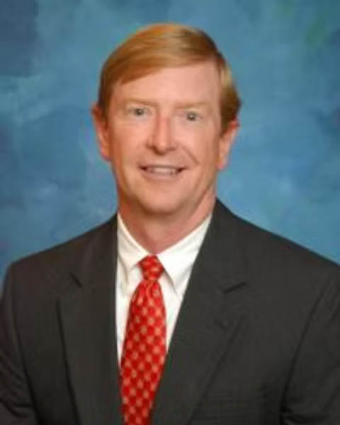 A professional headshot of a of a man with red hair wearing a dark suit, white shirt, and red tie with a patterned design, against a blue background.