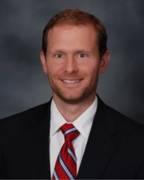 A professional headshot of a of a man with short red hair, wearing a black suit, white shirt, and red striped tie, smiling against a dark blurred background.