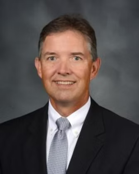 A professional headshot of a of a man in a suit with a white shirt and gray tie, smiling, against a gray background.