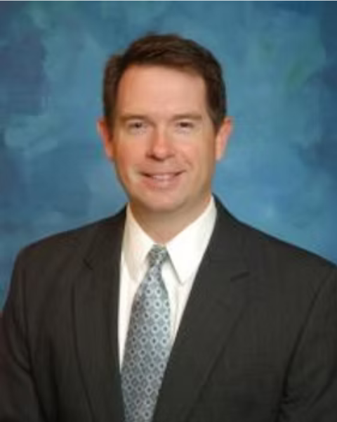 Professional headshot of a man in a dark suit, white shirt, and patterned tie, smiling in front of a blue textured background.