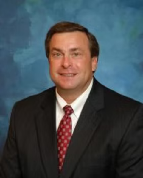 A professional headshot of a man in a black suit with a red patterned tie, smiling, against a blue backdrop.