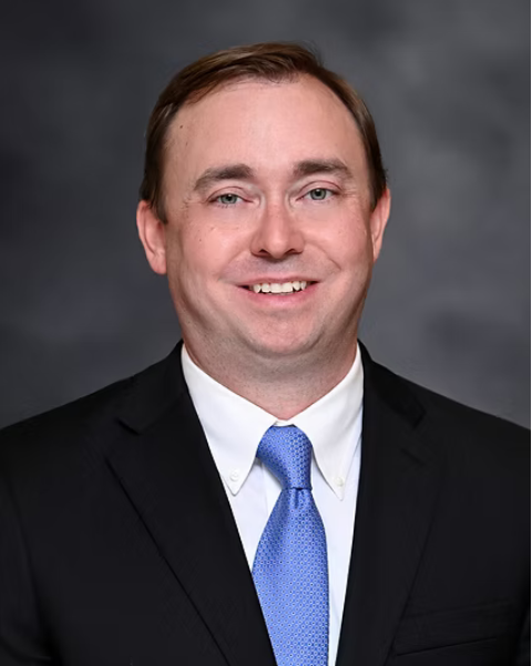 A professional headshot of a smiling man in a suit and blue tie against a gray background.