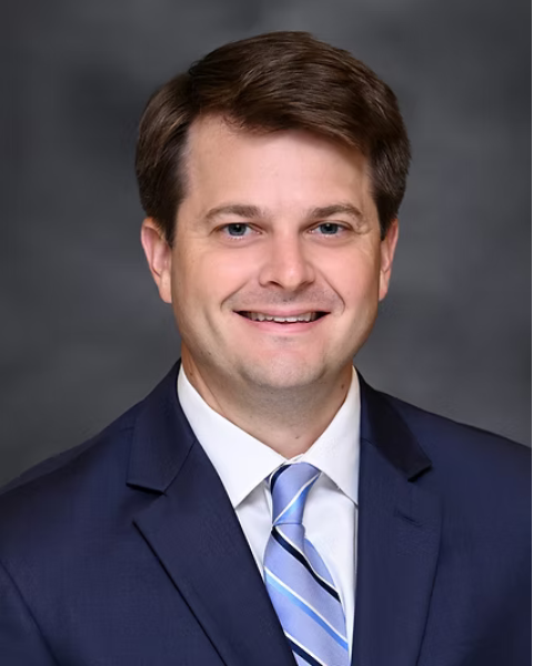 A professional headshot of a of a smiling man in a navy suit, white shirt, and light blue striped tie against a gray background.