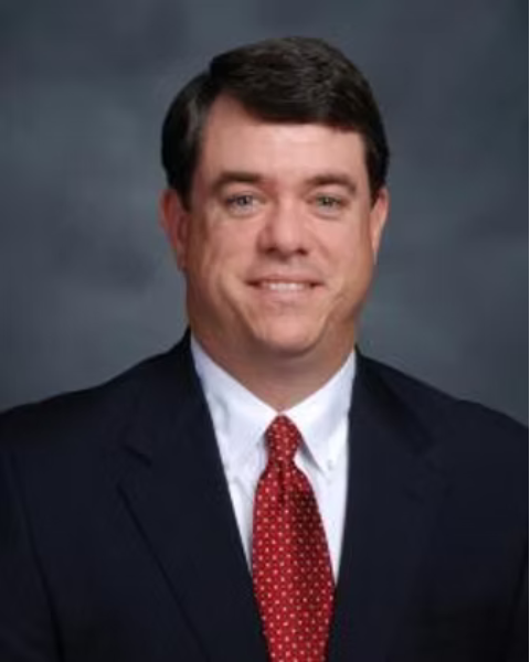 A professional headshot of a man with short dark hair, wearing a navy suit, white shirt, and red polka dot tie, against a gray background.