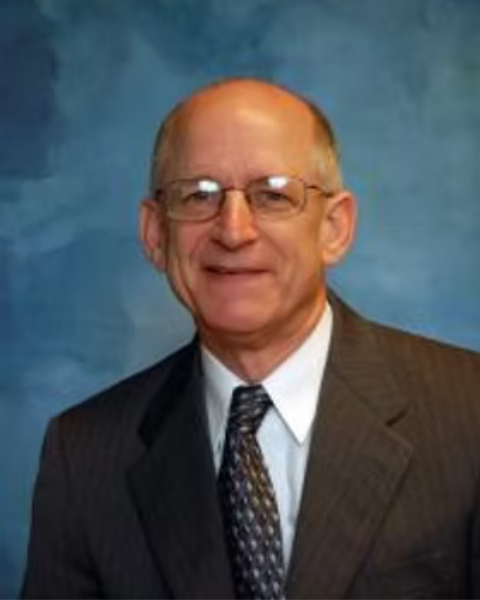 A professional headshot of a man wearing glasses, a dark pinstripe suit, a white shirt, and a patterned tie, smiling in front of a blue background.