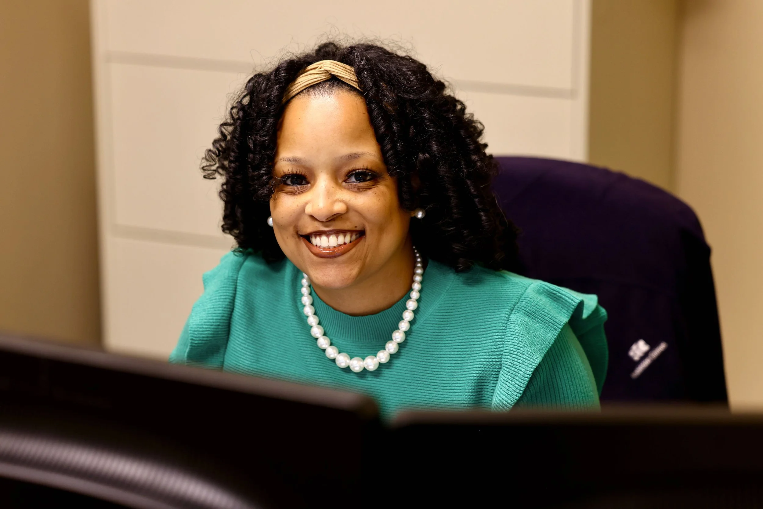 A smiling woman with curly hair, wearing a green top, pearl necklace, and earrings, sitting in an office.