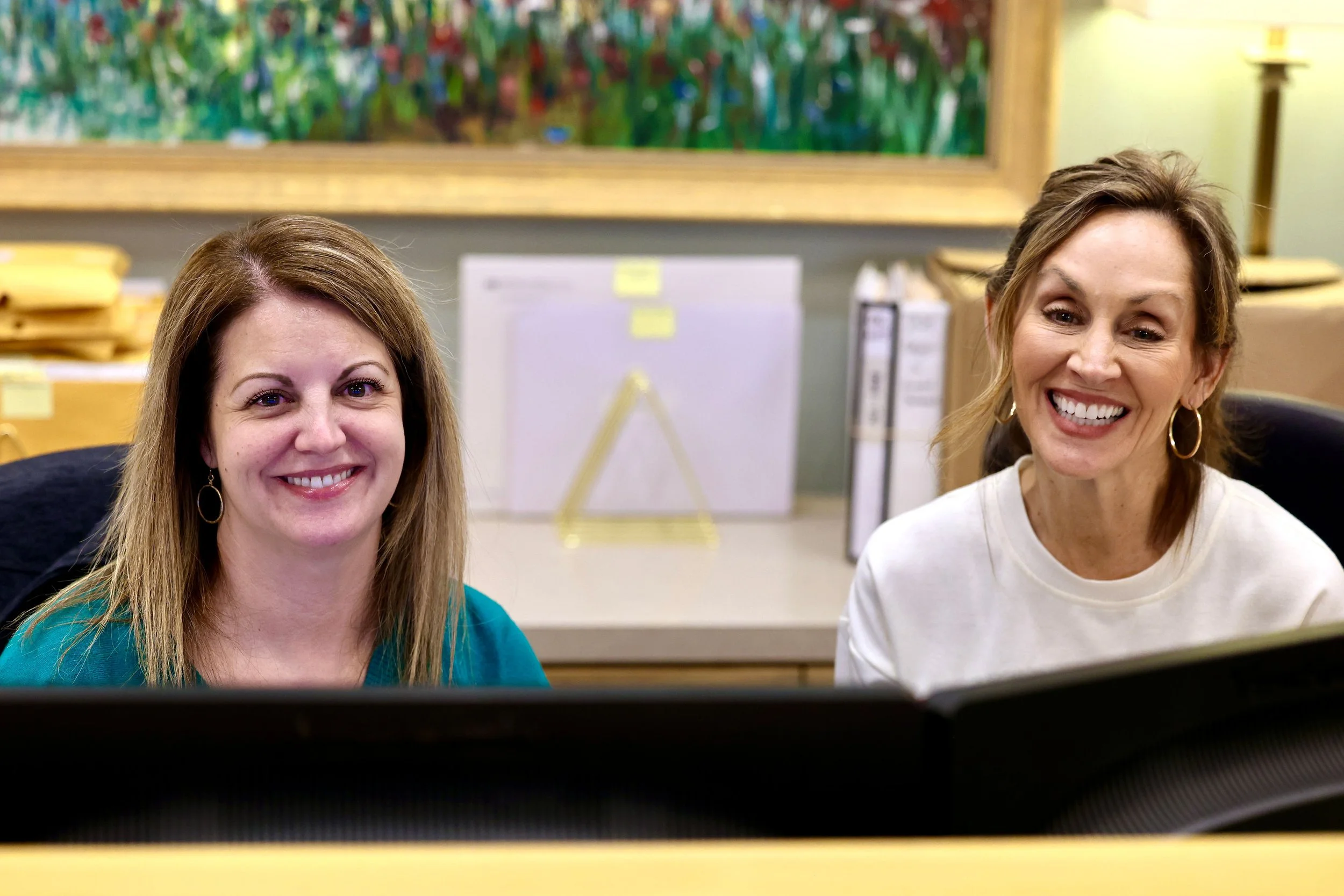 Two women sitting at a desk in an office, smiling and facing a computer monitor. Behind them are office supplies and a colorful abstract painting on the wall.