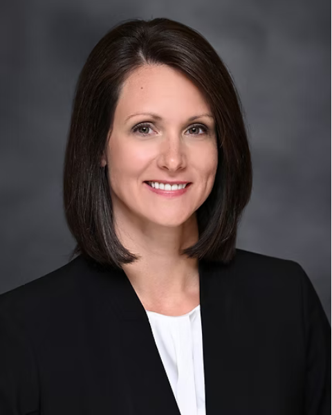 A professional headshot of a of a woman with shoulder-length brown hair, wearing a black blazer and white top, smiling against a gray background.